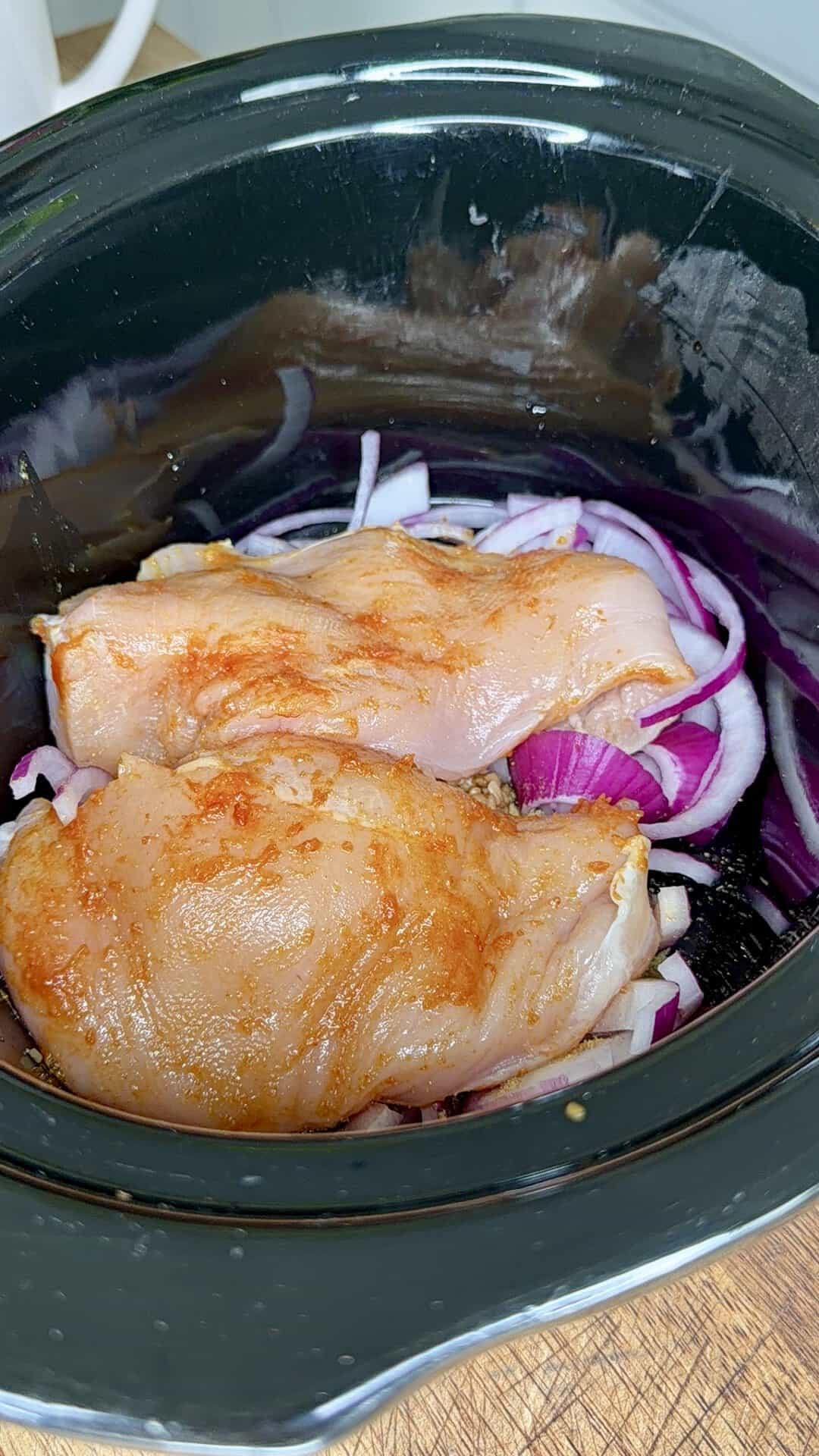 Raw, seasoned chicken breasts placed on a bed of sliced red onions inside a black slow cooker, ready to be cooked.