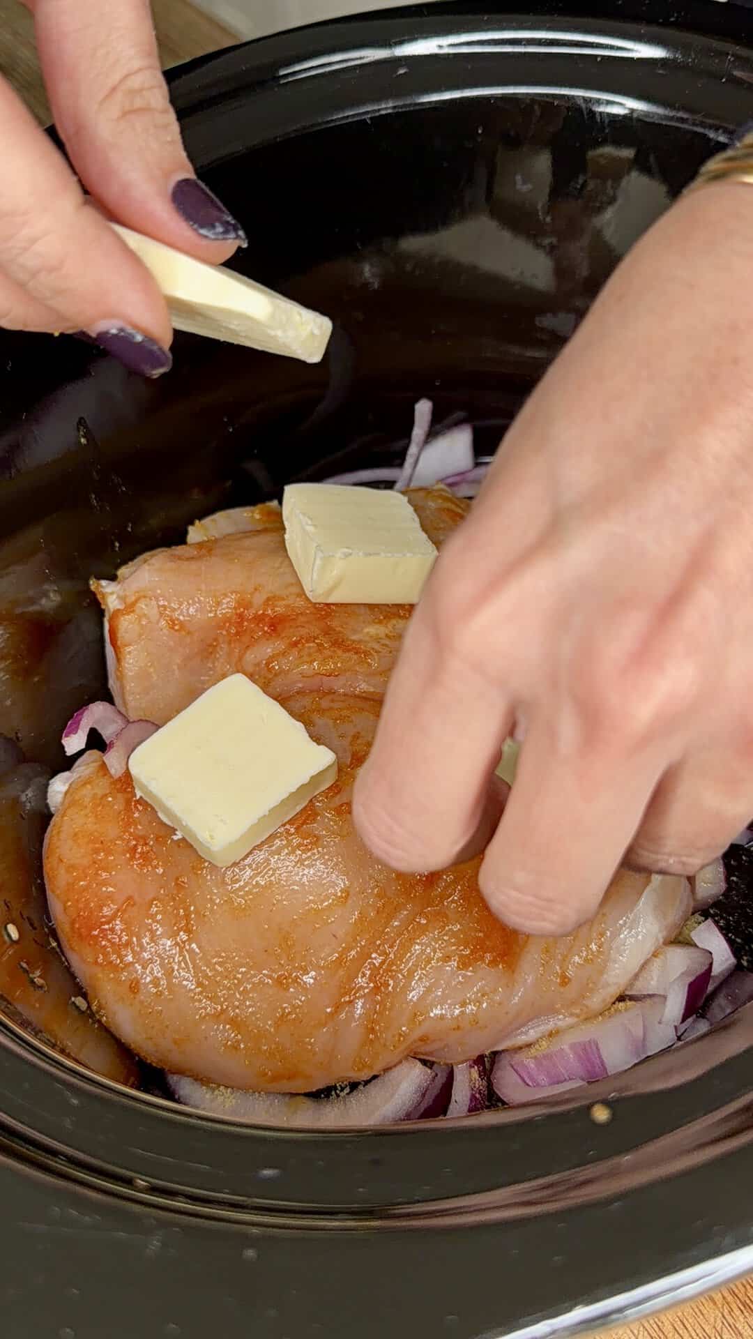 Two hands add pats of butter to seasoned raw chicken breasts placed on chopped red onions inside a black slow cooker.