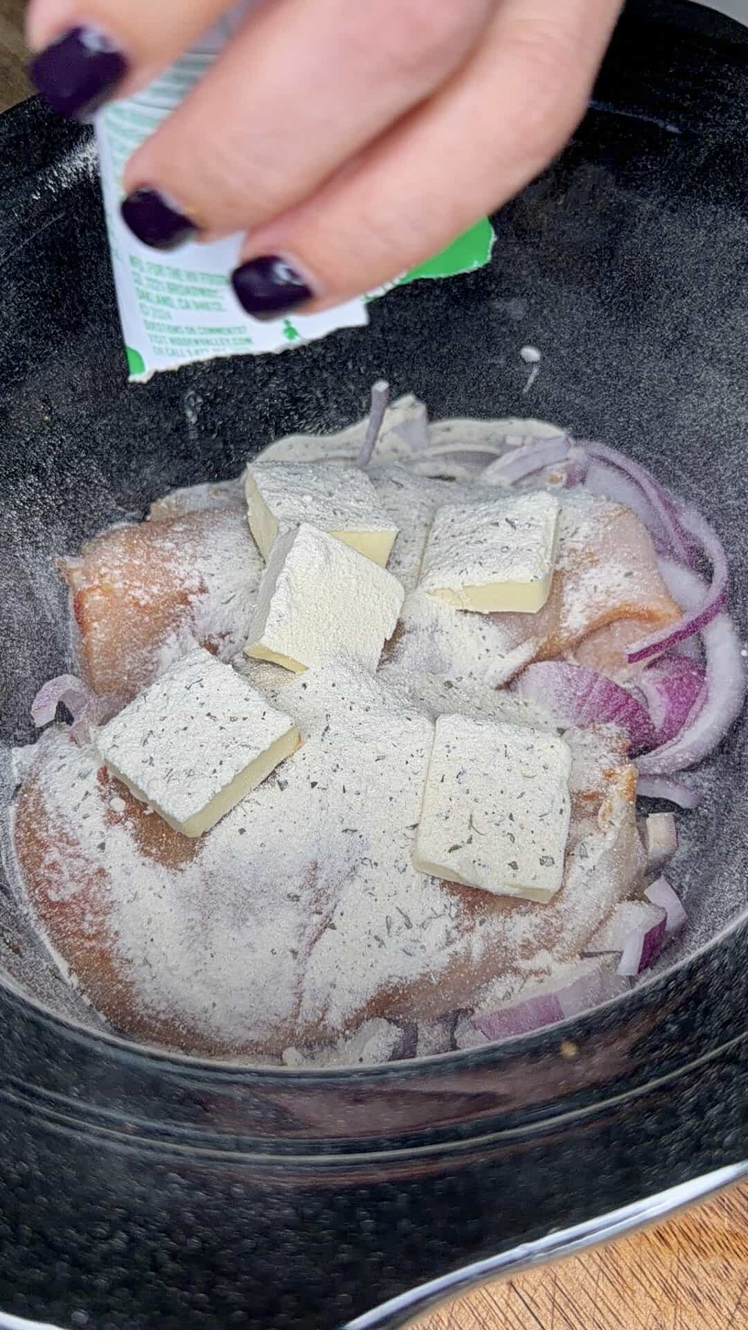 A hand sprinkles seasoning over raw chicken topped with sliced butter and chopped onions in a slow cooker.