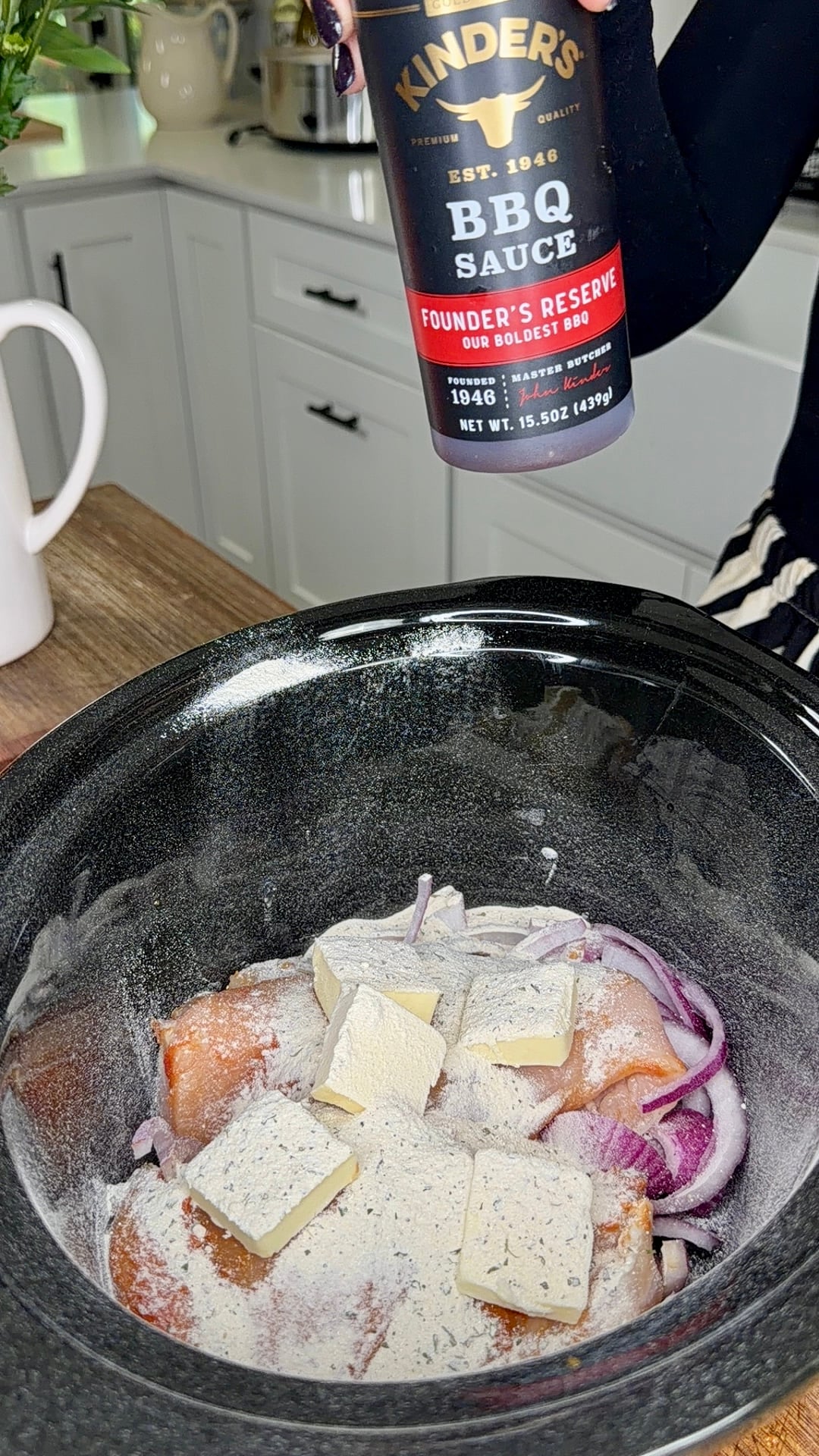 Raw chicken, sliced onions, seasoning, and butter pats in a slow cooker, with a person holding a bottle of Kinders BBQ Sauce above, ready to pour it in. White cabinets and kitchen items are in the background.