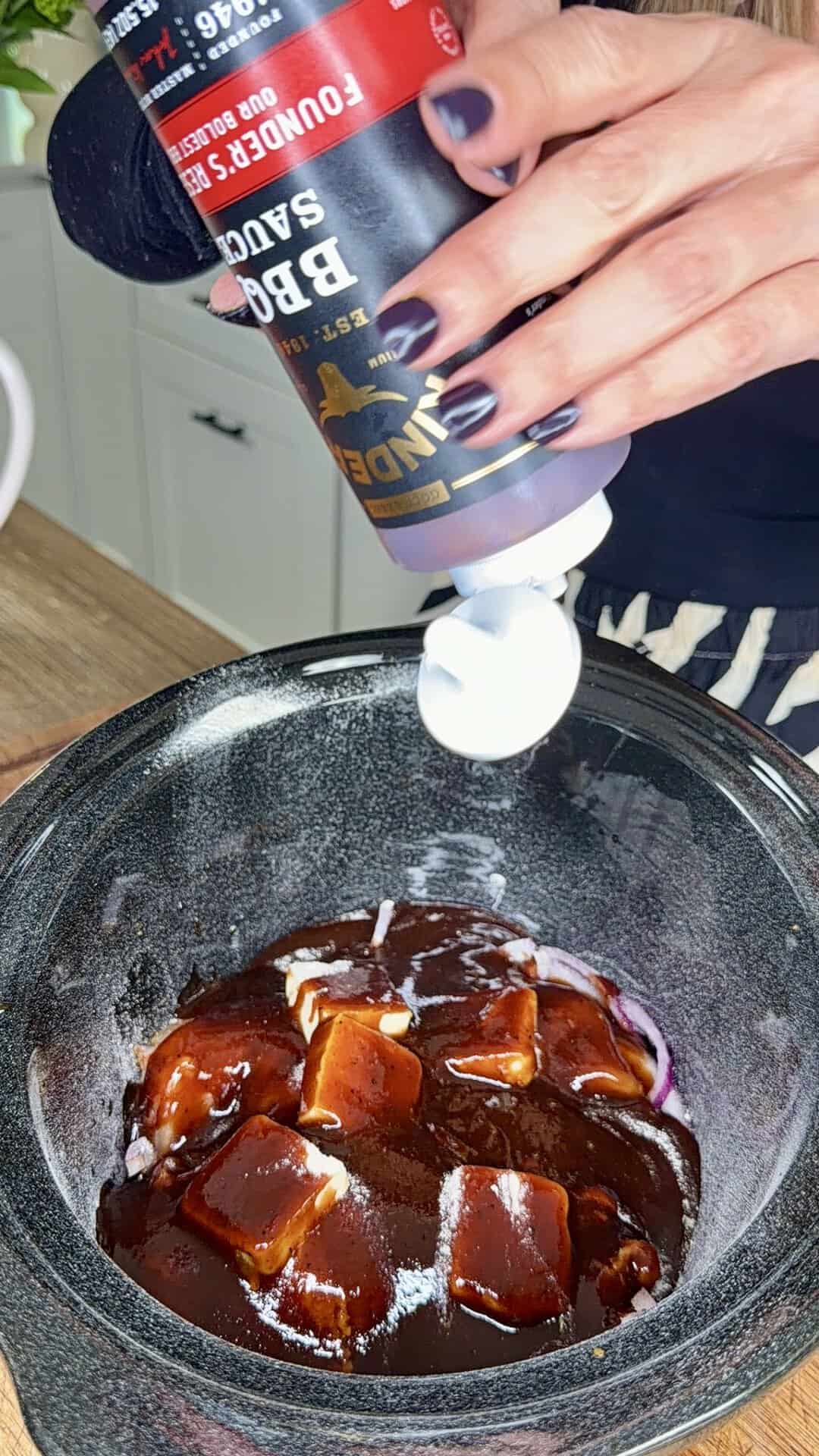 A hand pours BBQ sauce from a bottle into a slow cooker filled with cubed butter and sauce.