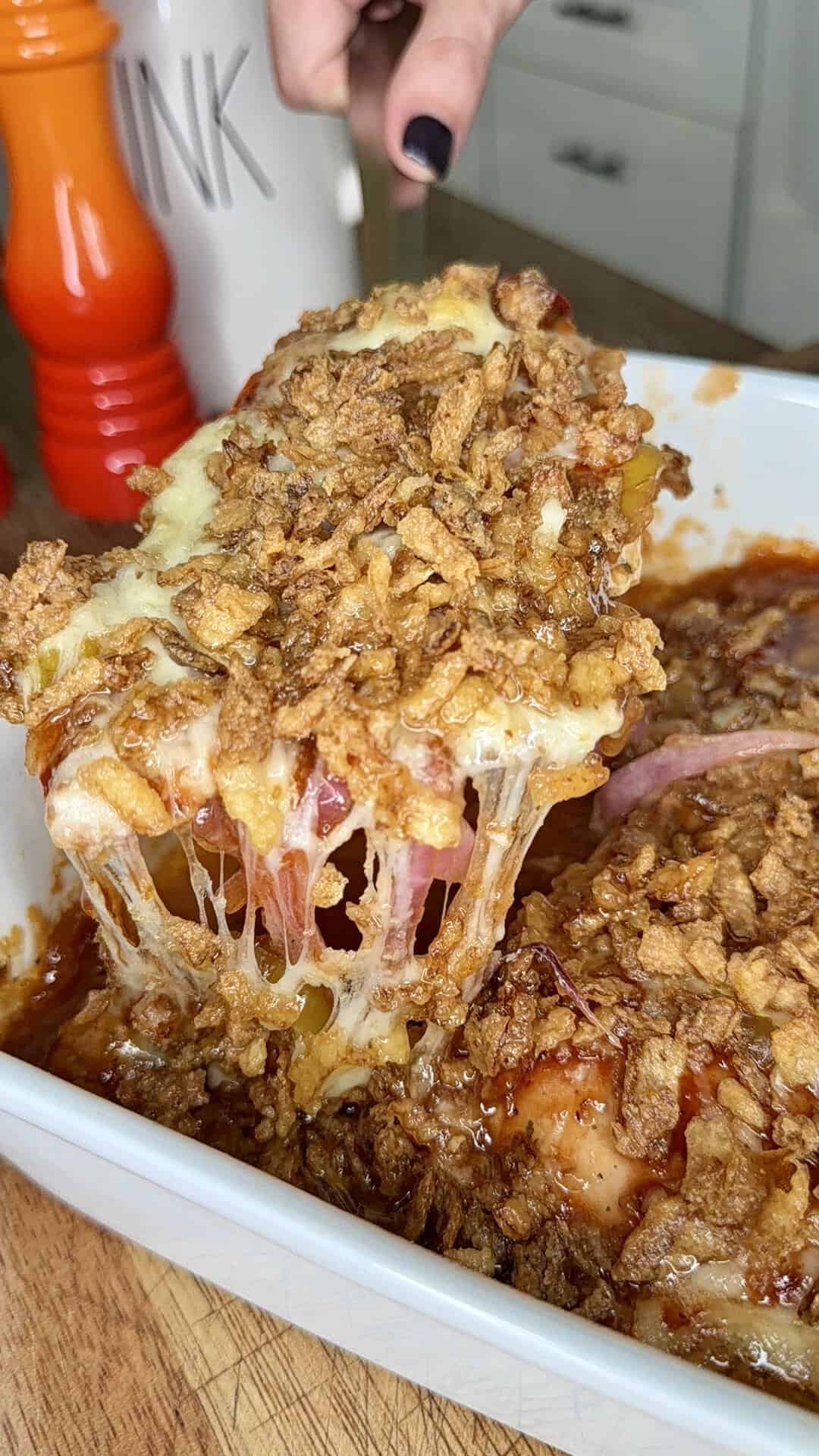 A hand lifts a serving of cheesy, crispy onion-topped casserole from a white baking dish, with melted cheese stretching and a red pepper grinder visible in the background.