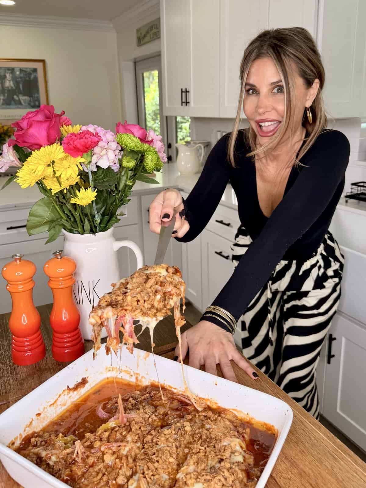 A woman in a black top and striped pants smiles while serving cheesy baked pasta from a white dish in a bright kitchen with a vase of colorful flowers and orange salt and pepper shakers nearby.