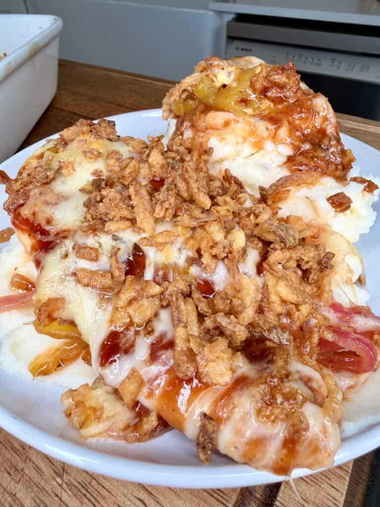 Close-up of a plate with cheesy, baked casserole topped with crispy fried onions, melted cheese, and visible layers of sauce and vegetables. The dish is set on a wooden counter in a kitchen.
