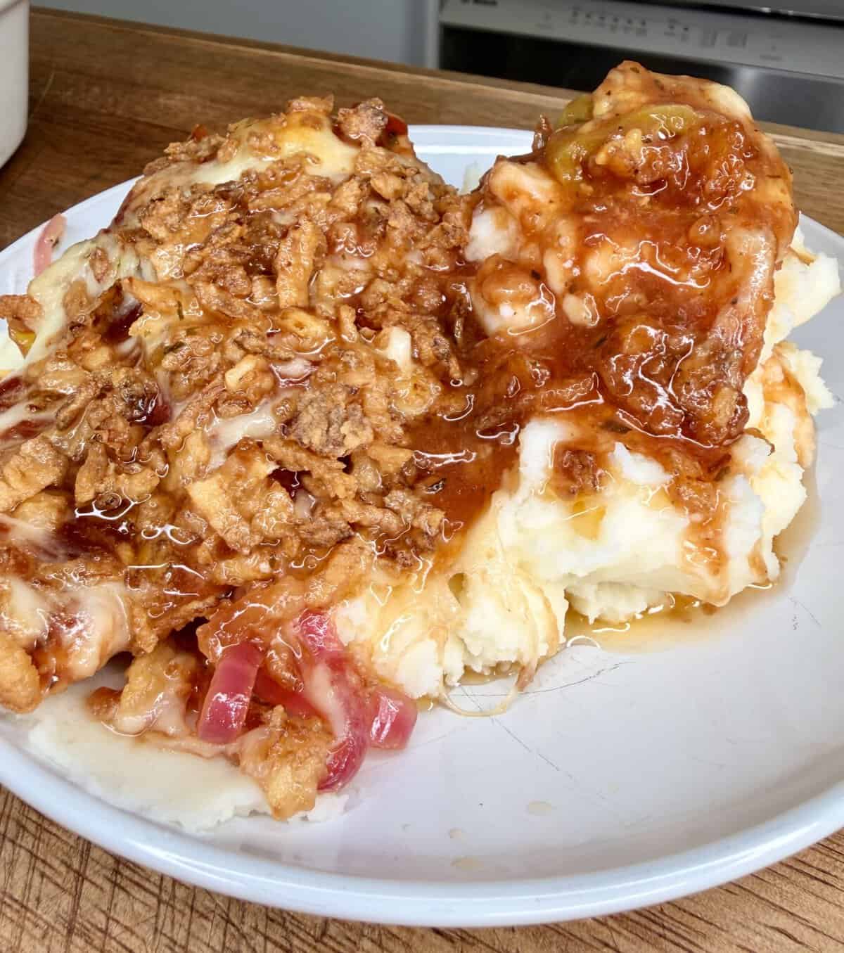 Mashed potatoes topped with crispy fried onions, shredded cheese, red onions, and smothered in brown gravy, served on a white plate on a wooden surface.