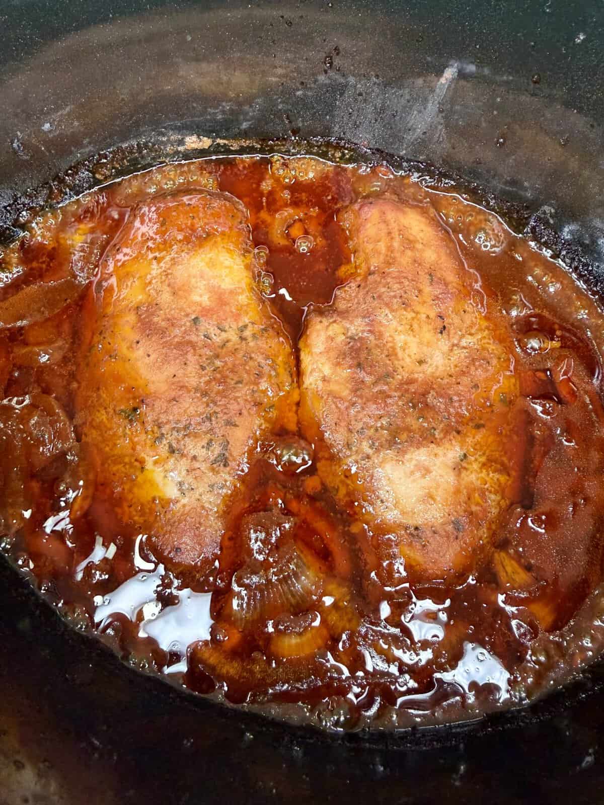 Two seasoned chicken breasts are simmering in a rich, reddish-brown sauce with visible chunks of onion in a black slow cooker or pot.