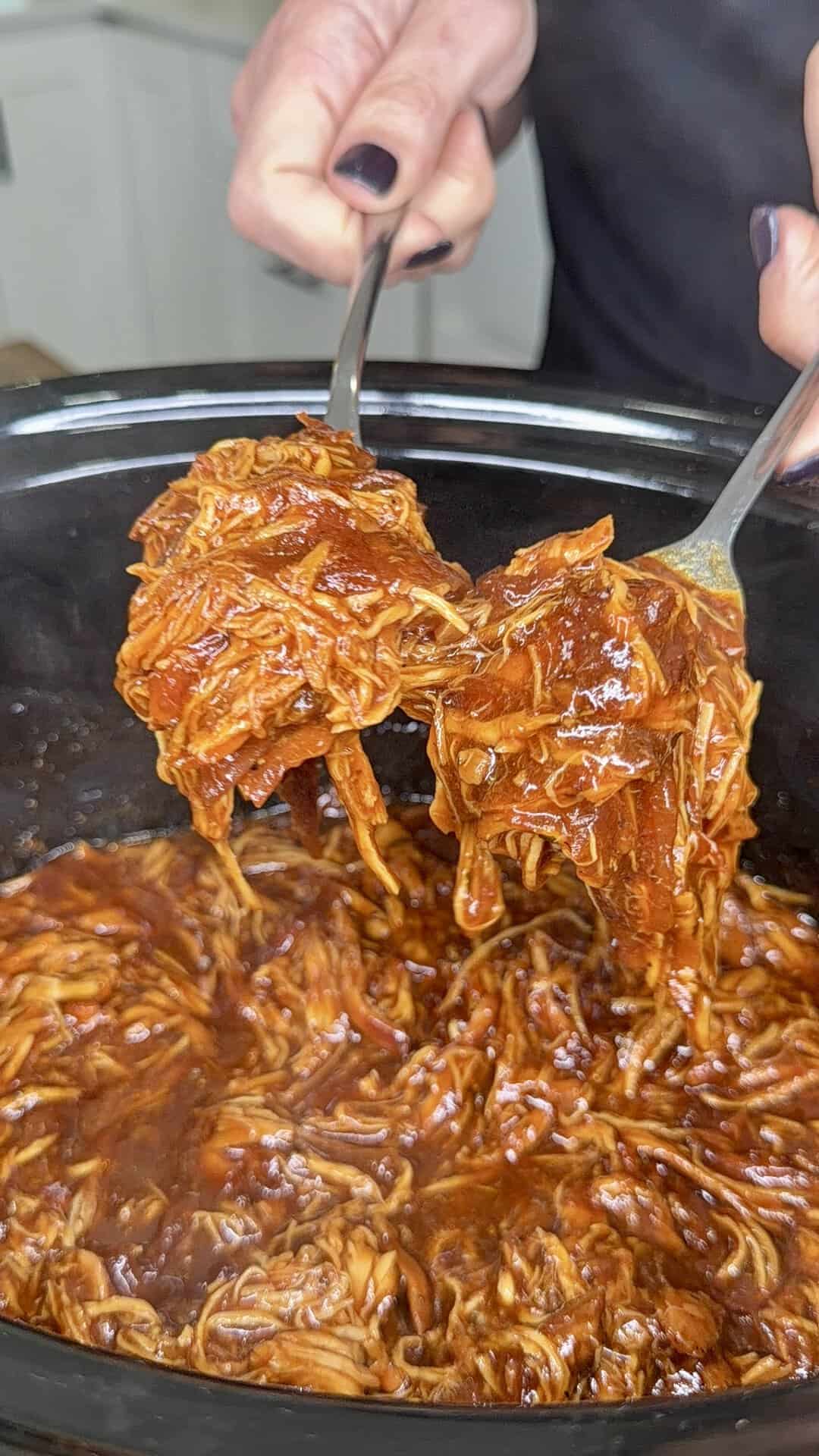 Two forks lift saucy shredded barbecue chicken from a slow cooker. A persons hands with dark nail polish hold the forks over the steaming, rich, reddish-brown mixture.