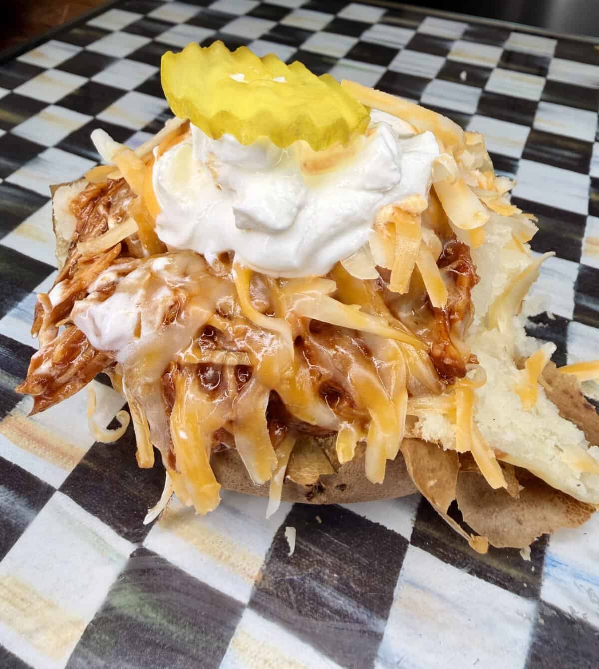 A baked potato topped with shredded cheese, barbecue pulled meat, sour cream, and two pickle slices, served on a black and white checkered surface.