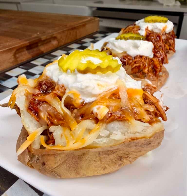 A baked potato topped with shredded barbecue chicken, melted cheese, onions, sour cream, and pickle slices, served on a white plate.