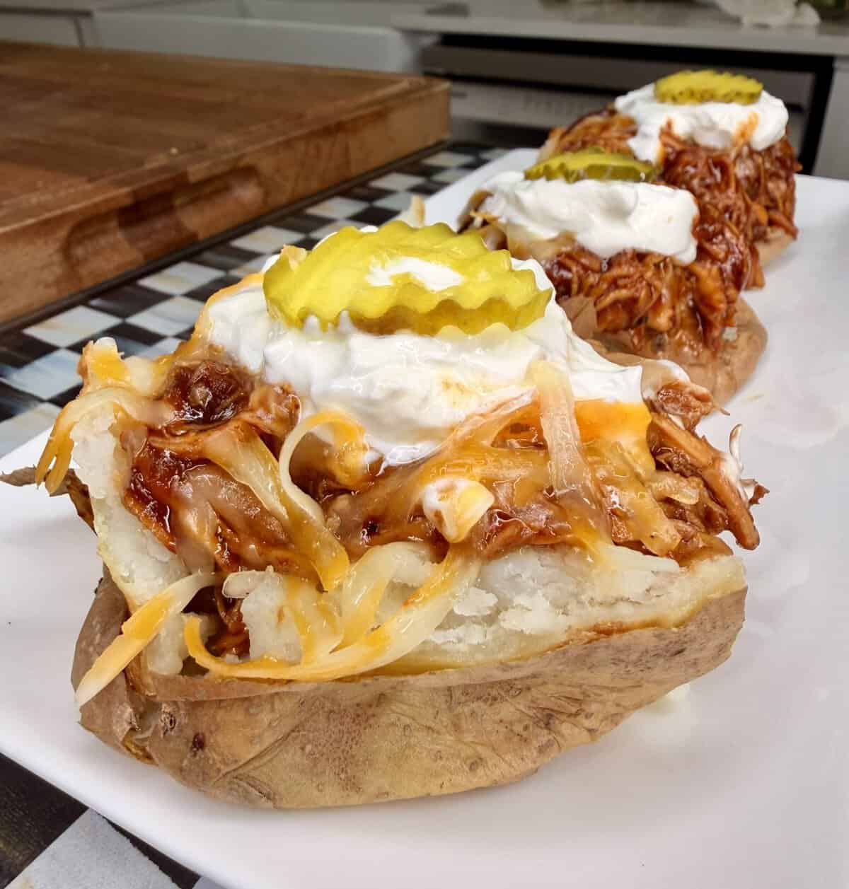 A baked potato topped with shredded barbecue chicken, melted cheese, onions, sour cream, and pickle slices, served on a white plate.