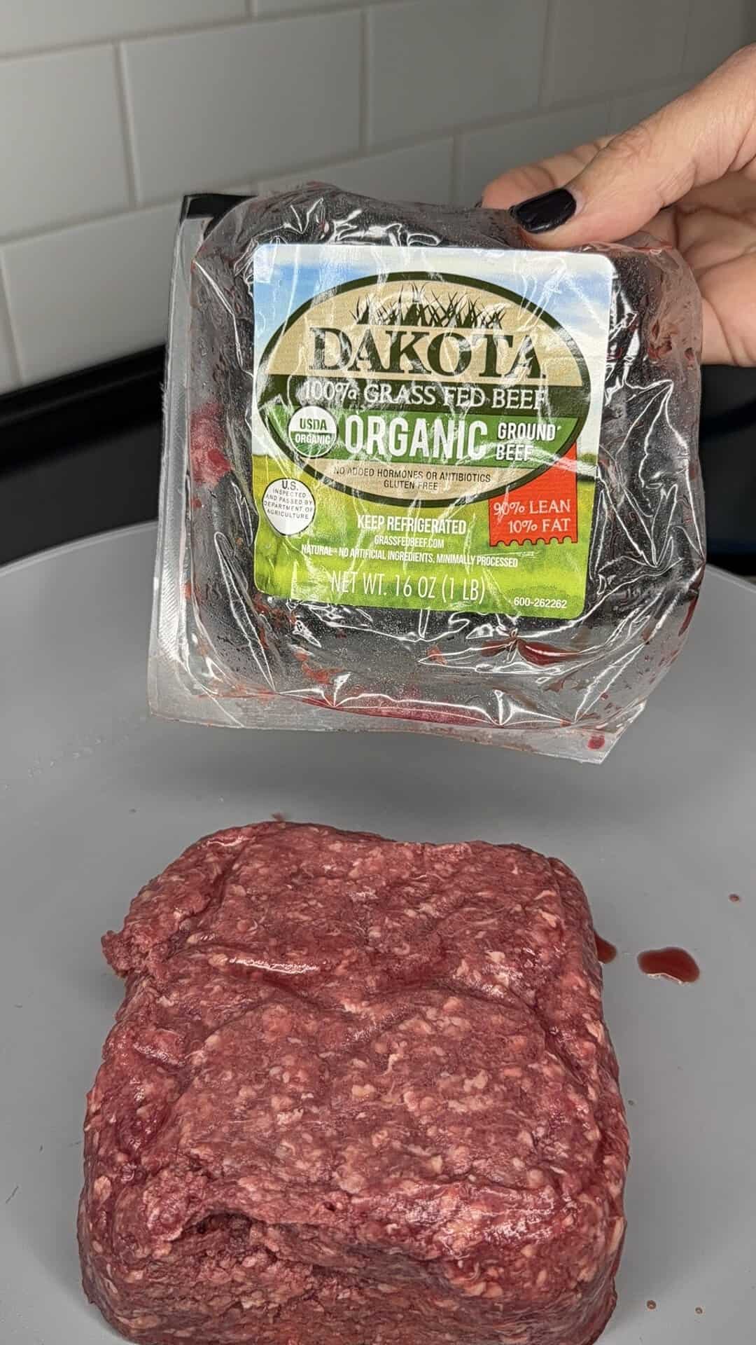A hand holds a package of organic grass-fed ground beef above a portion of raw ground beef on a counter, with a white tiled wall in the background.