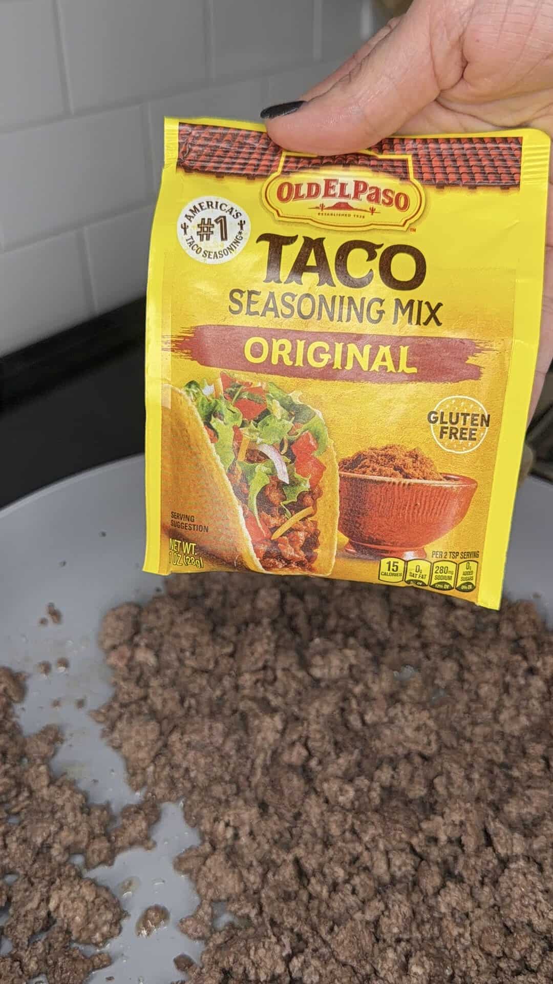 A hand holds a packet of Old El Paso Taco Seasoning Mix above a pan of cooked ground beef on a stove, ready to add flavor.