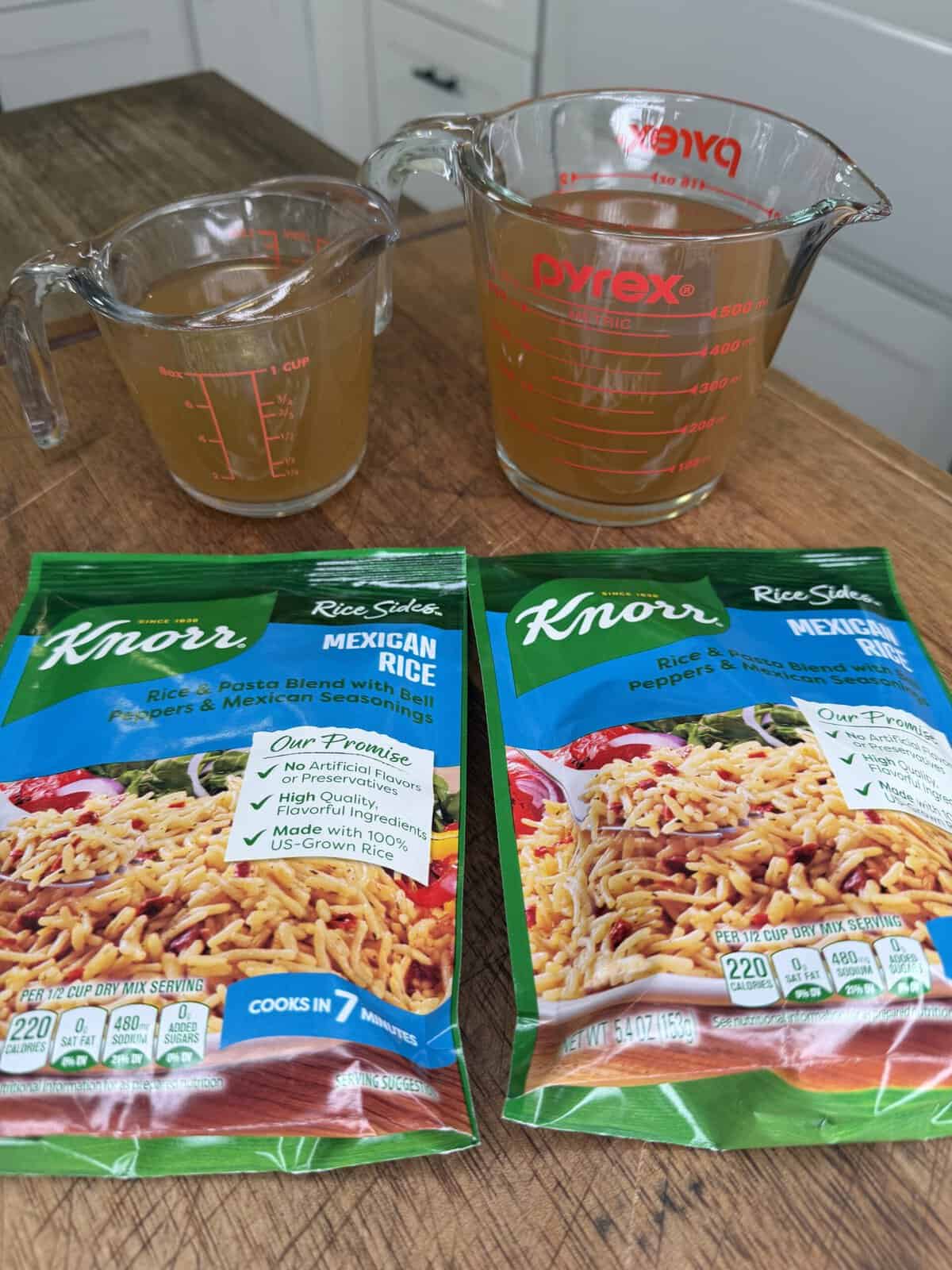 Two measuring cups with broth and water sit behind two Knorr Rice Sides Mexican Rice packets on a wooden kitchen counter, with white cabinets and drawers in the background.