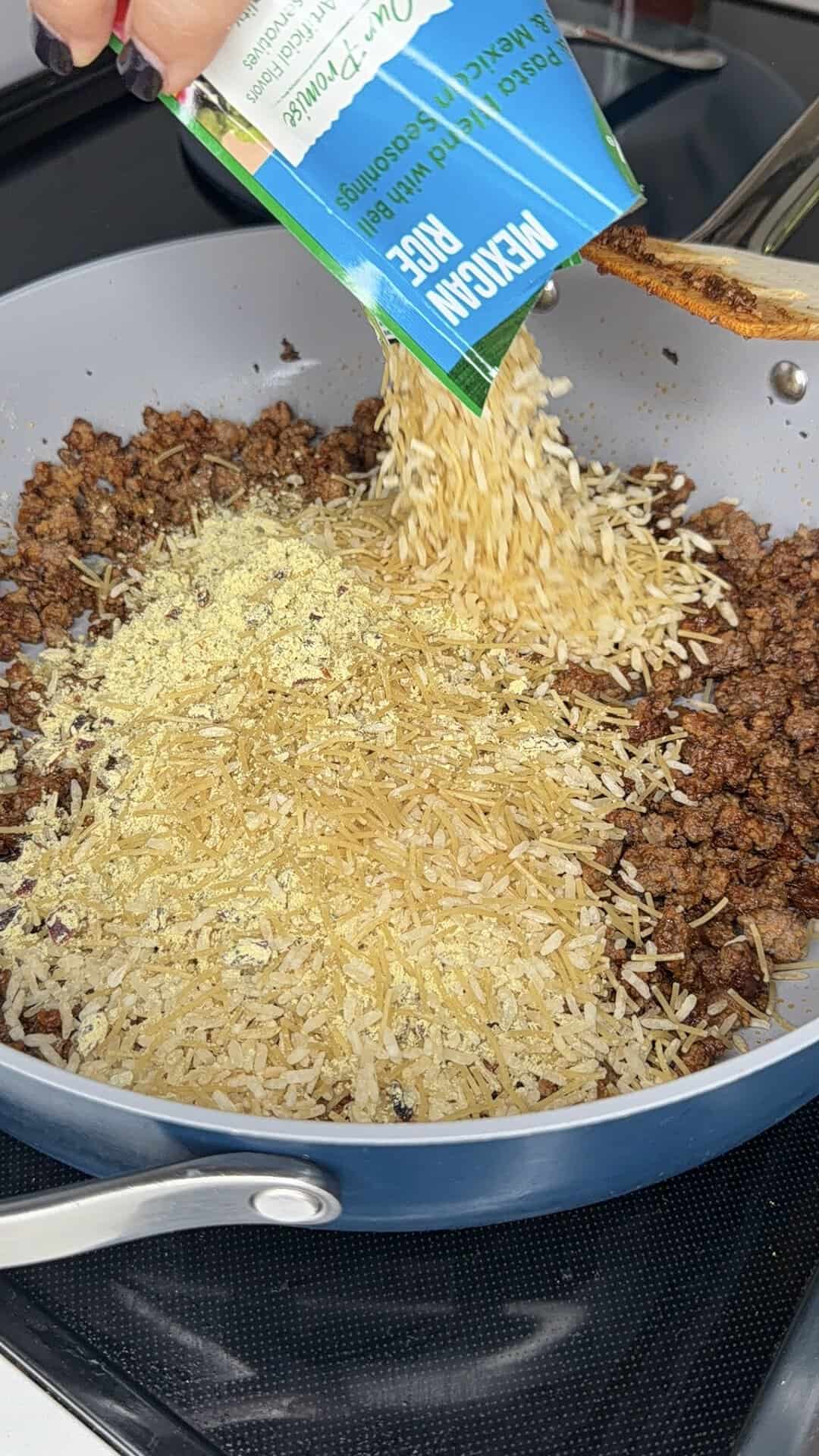 A hand pours uncooked rice from a box into a skillet with browned ground meat and seasonings, mixing the ingredients together while cooking on a stovetop.