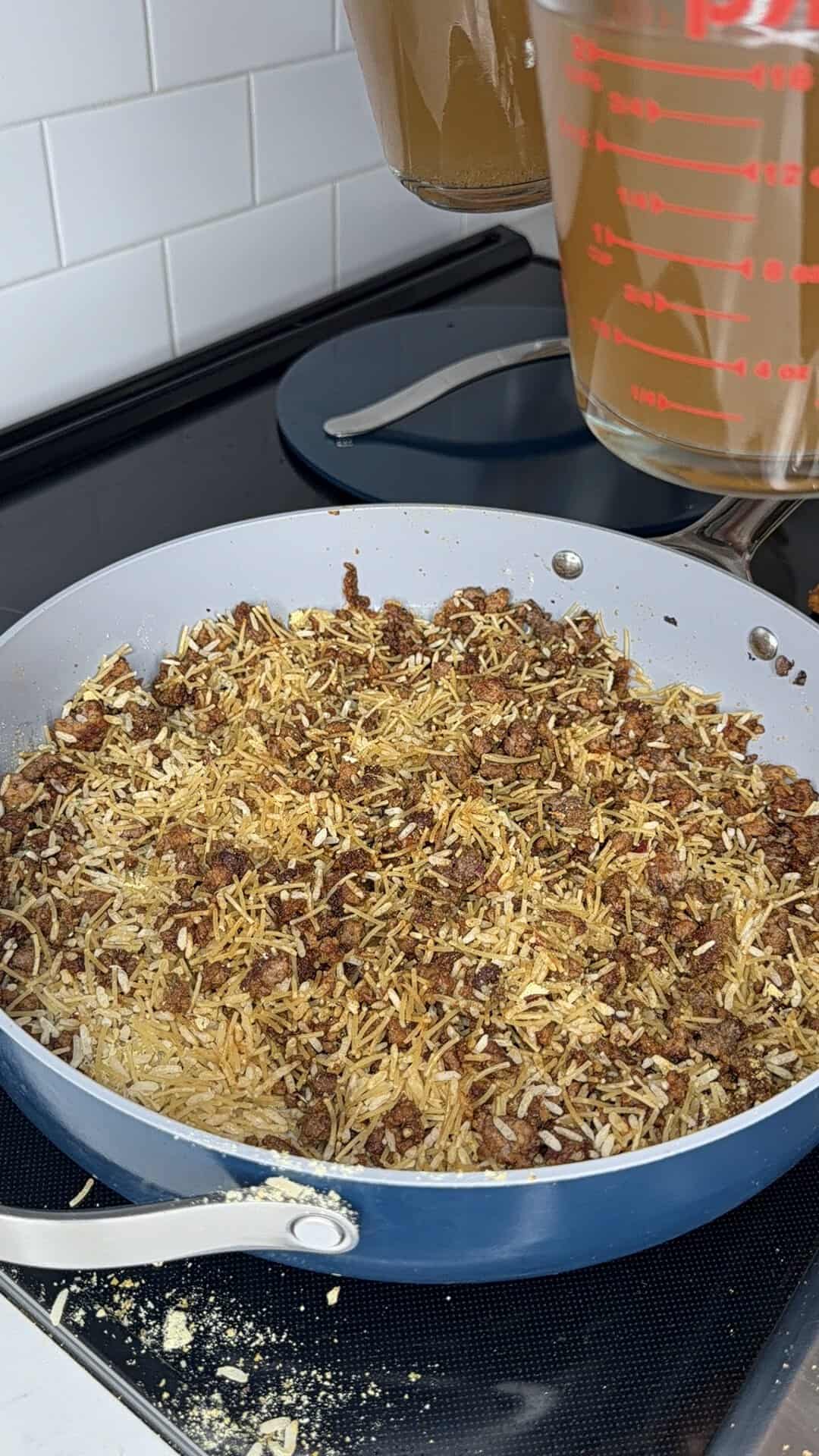 A blue frying pan on a stove with browned ground meat and uncooked rice. A hand pours liquid from a measuring cup above the pan. White subway tile backsplash is visible.