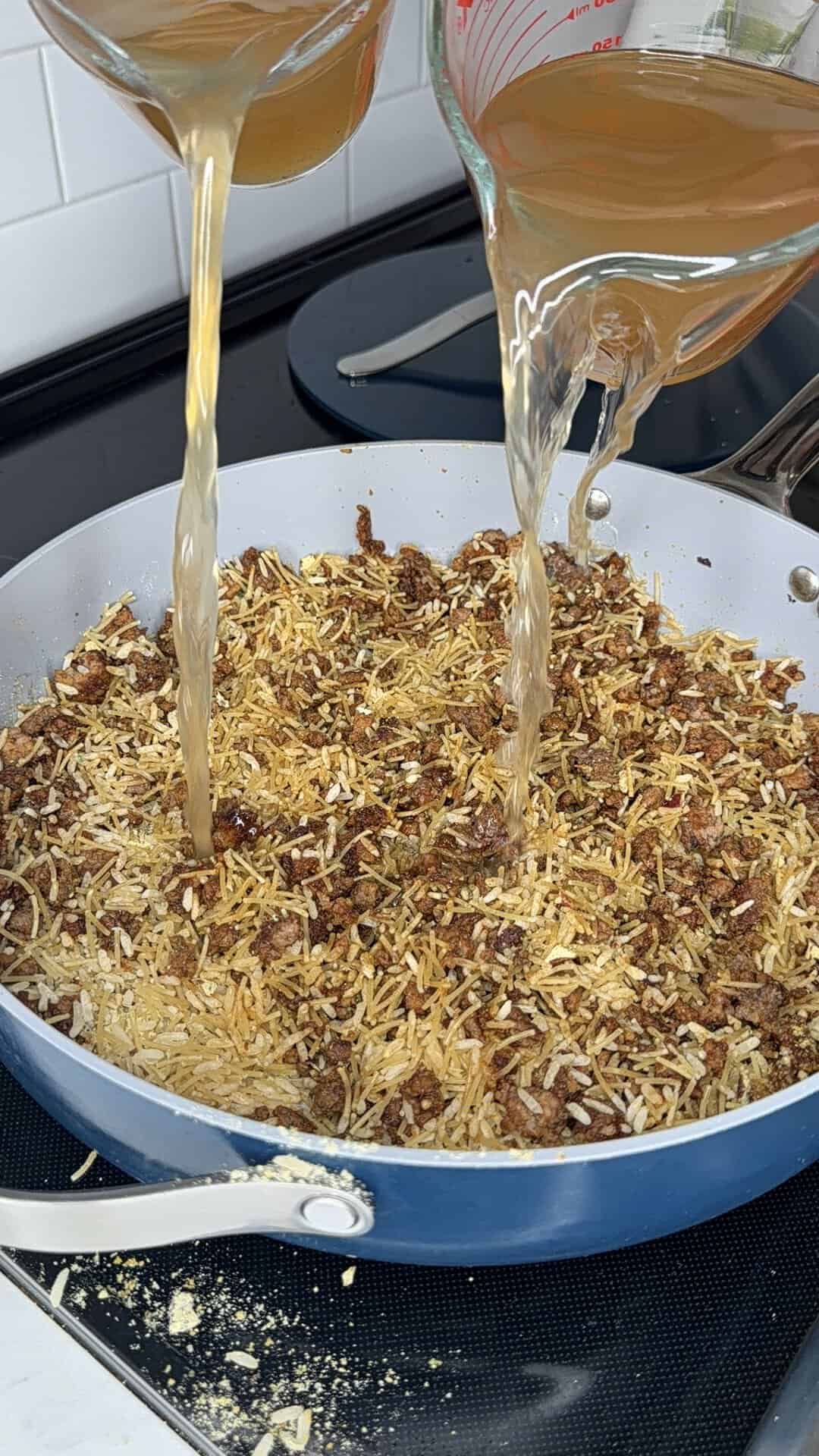 Beef and shredded potatoes cooking in a pan as two measuring cups pour broth over the mixture on a stovetop.