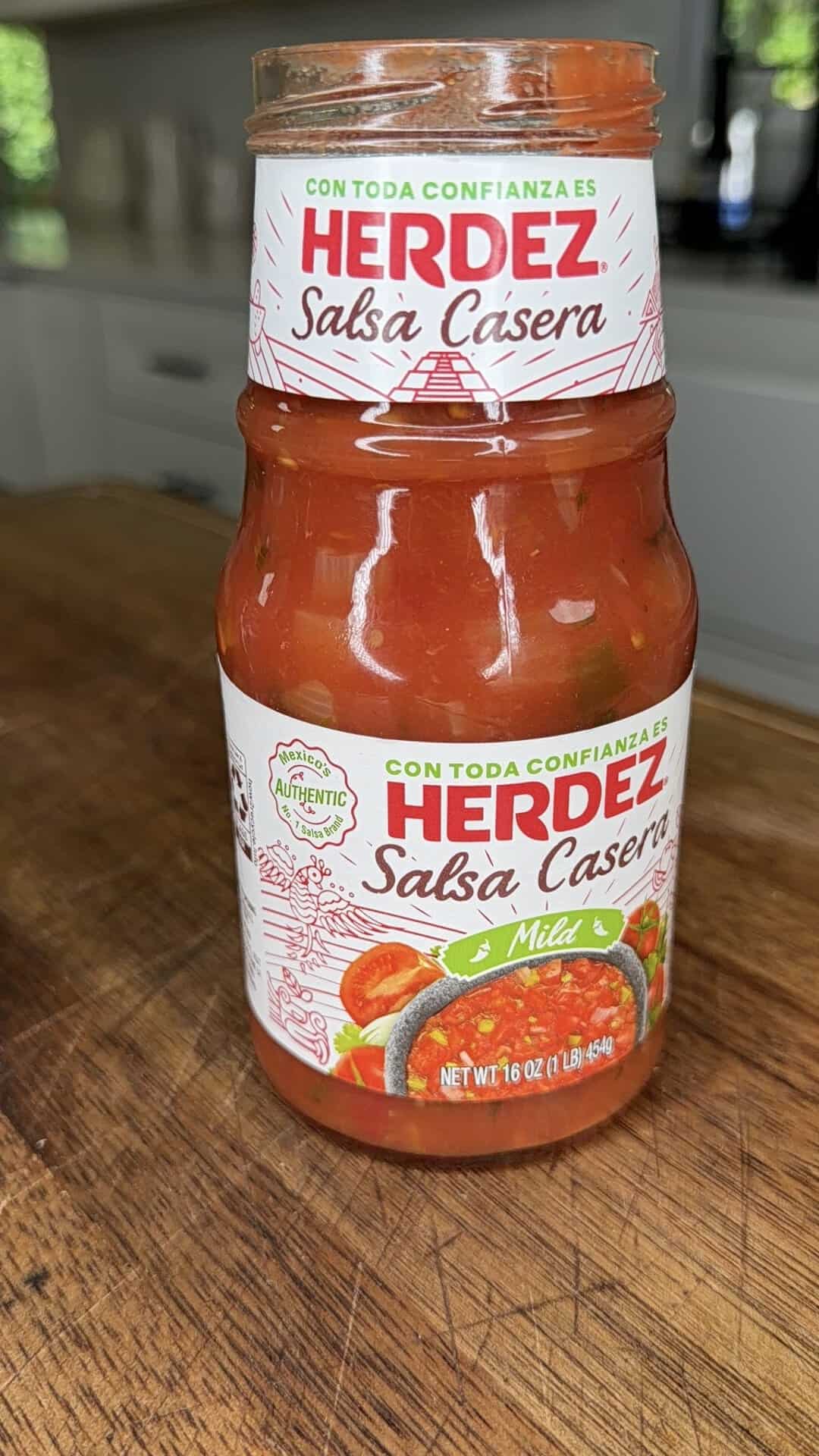 A jar of Herdez Salsa Casera Mild sits on a wooden countertop in a kitchen, displaying the label in both English and Spanish.