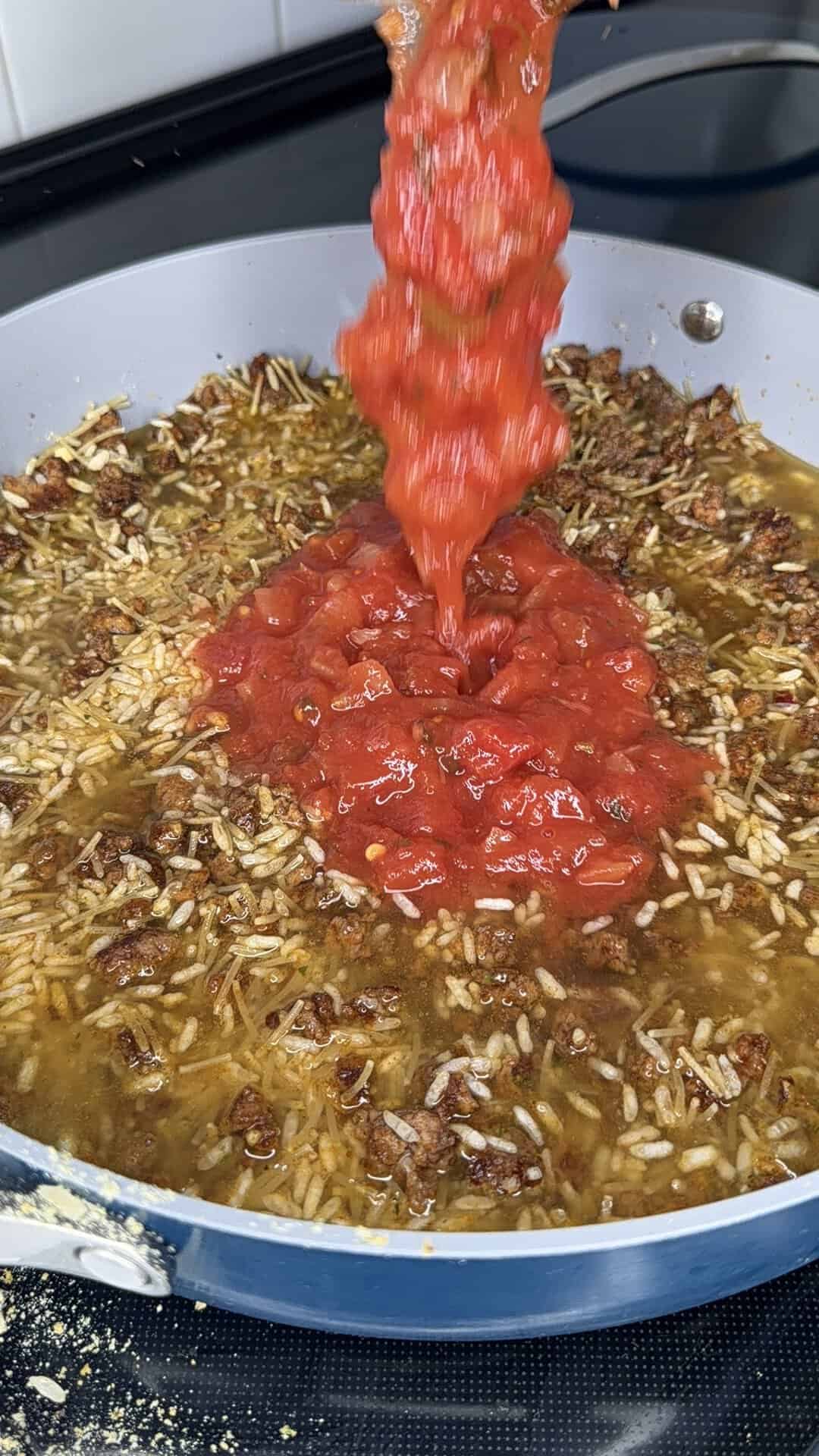Chopped tomatoes are being poured into a skillet containing cooked rice, ground meat, and broth, creating a hearty mixture on a stovetop.