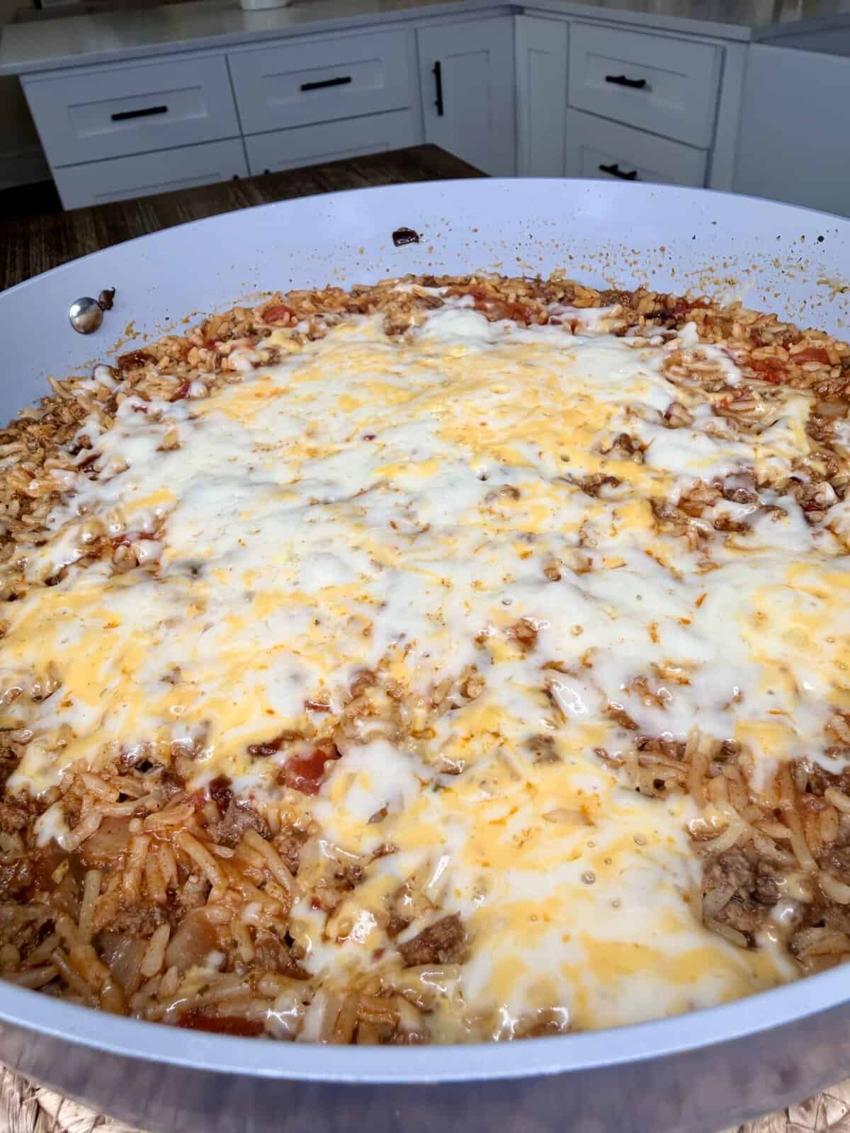 A large skillet filled with cheesy ground beef and rice casserole sits on a kitchen counter, melted cheese covering the top.