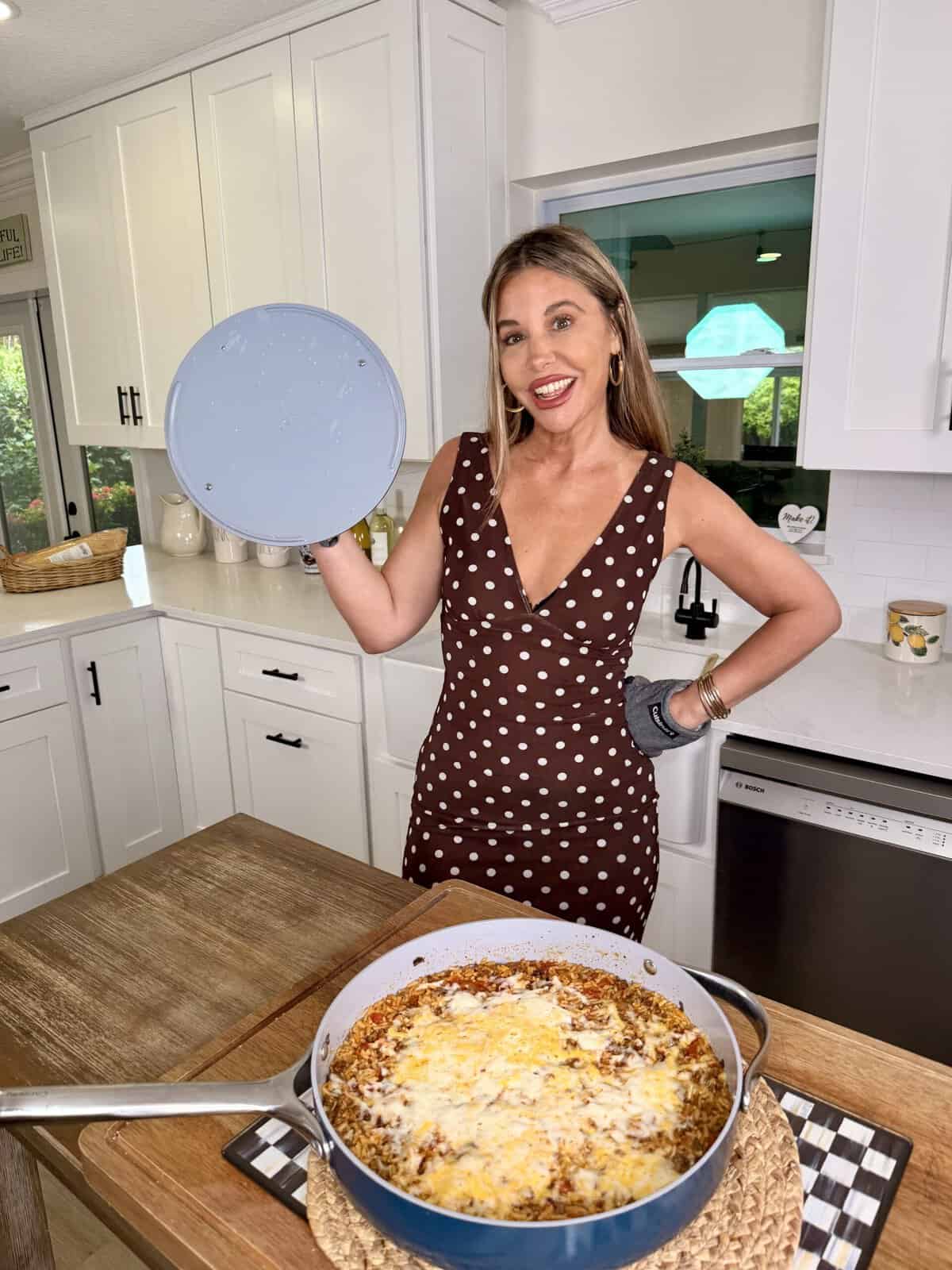 A woman in a brown polka dot dress stands in a kitchen, smiling and holding a pot lid. In front of her, theres a large baked pasta dish on the table.