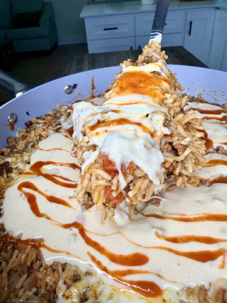 A close-up of a skillet filled with rice and meat, topped with creamy white sauce and drizzled with hot sauce. A large spatula lifts a serving from the skillet. A kitchen counter is visible in the background.