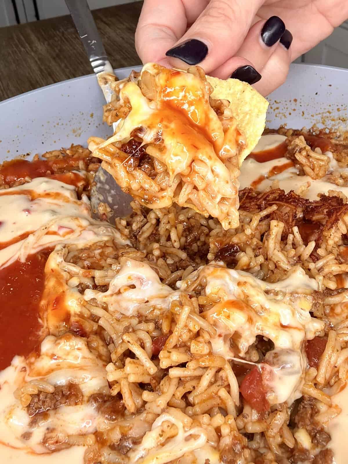 A hand with dark nails holds a chip dipped in cheesy rice, ground meat, and tomato salsa from a skillet full of the same mixture.