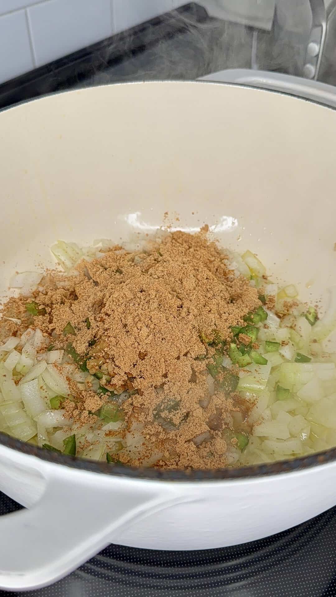 Chopped onions and green peppers are sautéing in a white pot, with a heap of brown seasoning or spice blend sprinkled on top. Steam rises as the ingredients cook.
