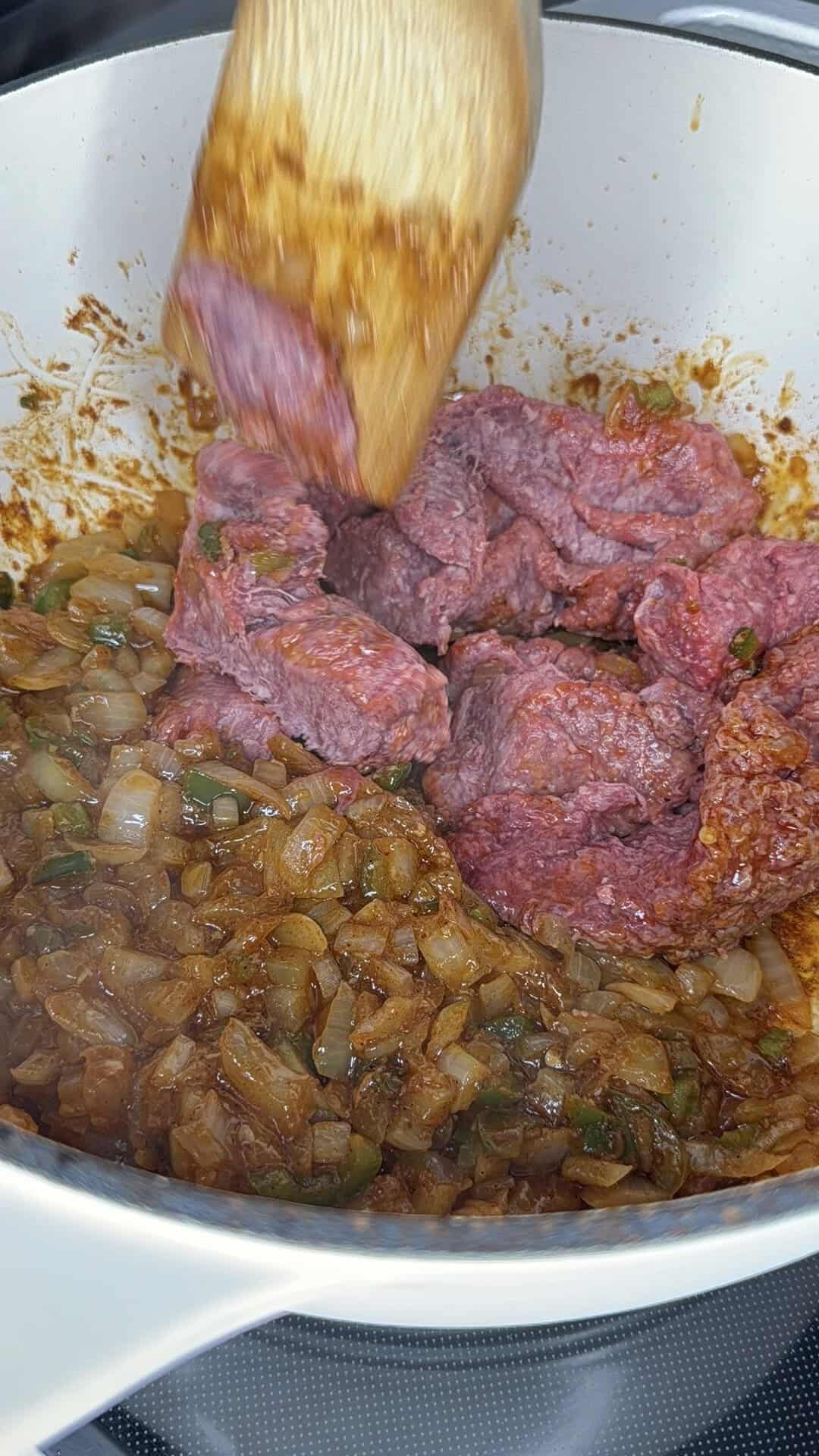 Chunks of raw beef being stirred into sautéed onions and spices in a white pot with a wooden spatula.