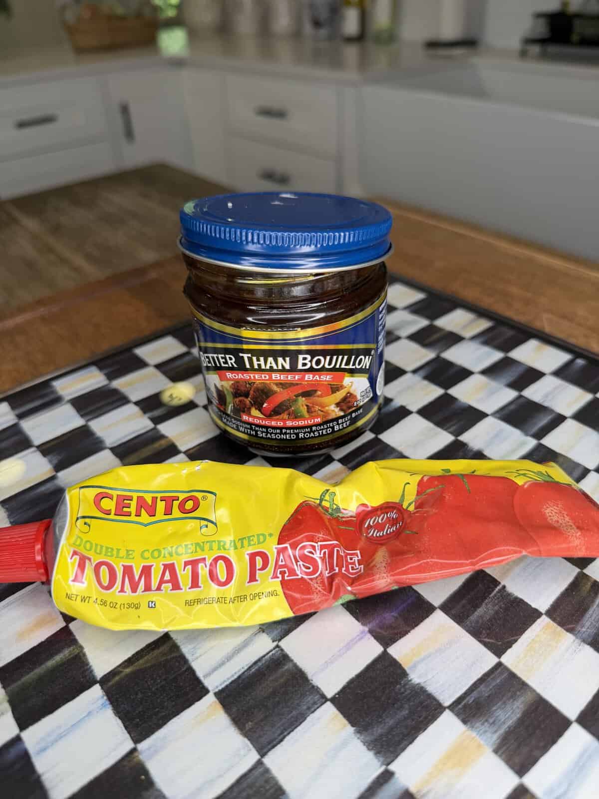 A jar of Better Than Bouillon Roasted Beef Base and a tube of Cento Tomato Paste sit on a black-and-white checkered surface in a kitchen.