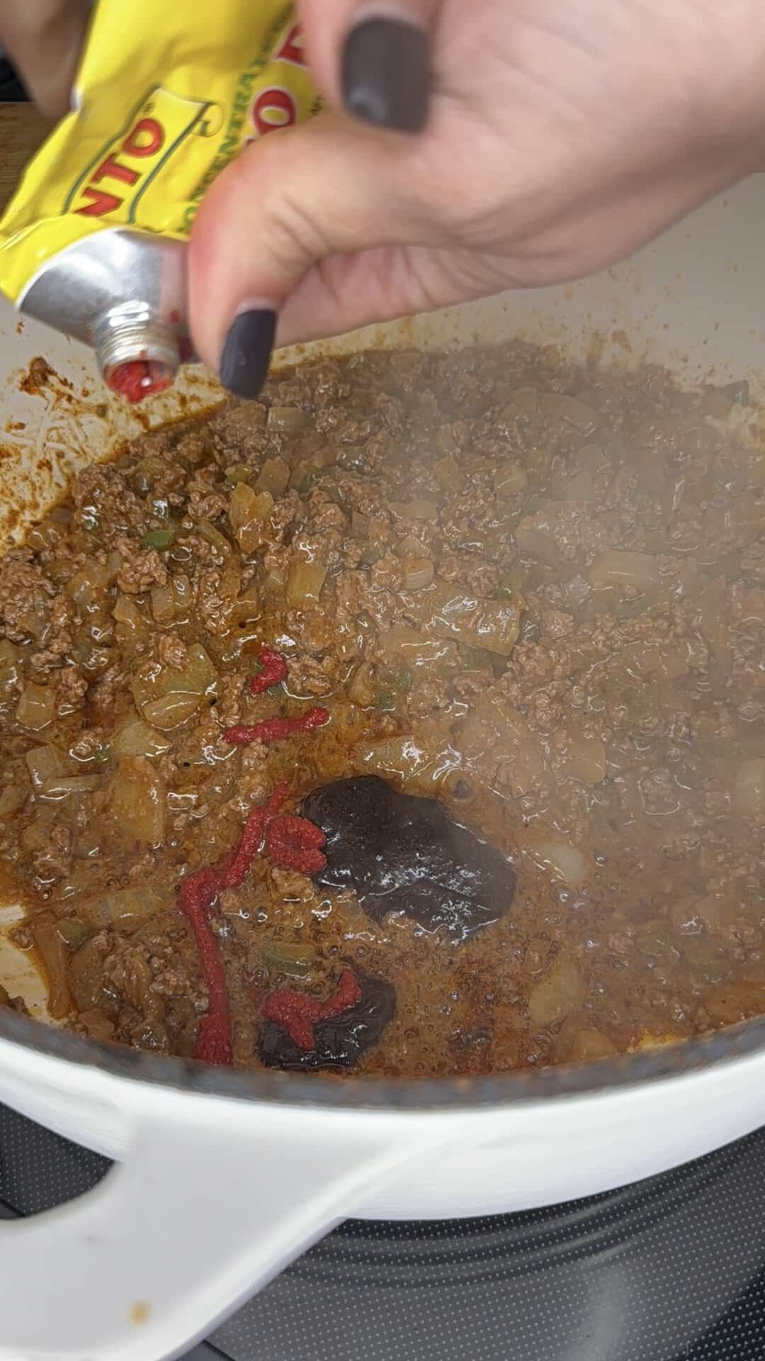 A hand squeezes tomato paste from a yellow tube into a pot of browned ground meat and onions on a stovetop. Steam rises from the mixture as it cooks.