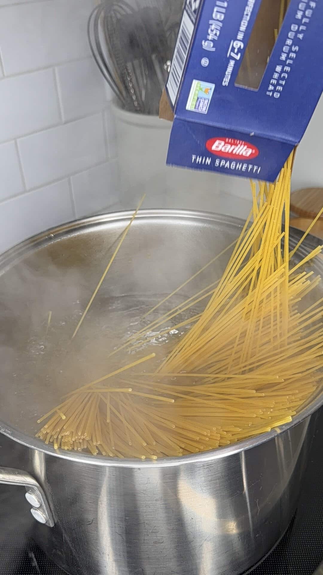 A box of Barilla thin spaghetti is being poured into a large pot of boiling water on a stove, with steam rising from the pot.