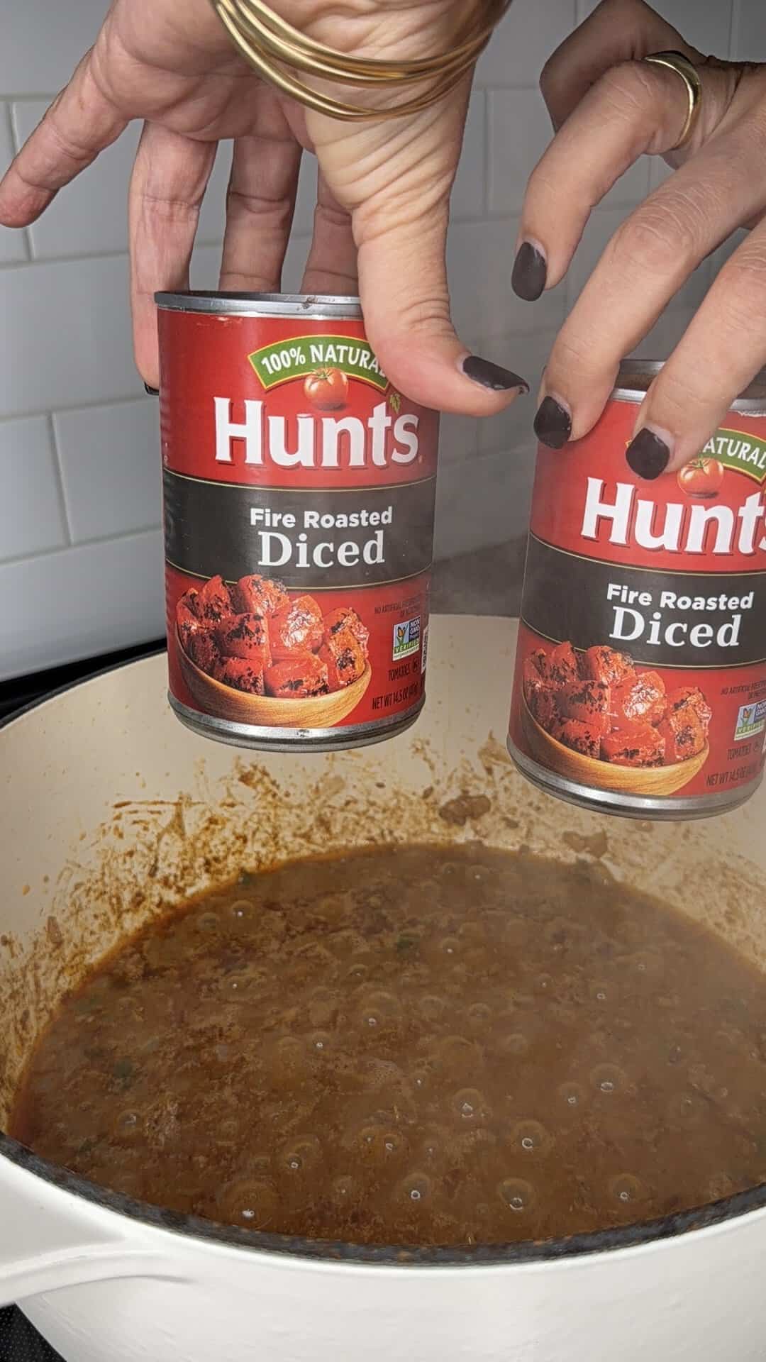 Two hands with dark nail polish hold cans of Hunts Fire Roasted Diced tomatoes above a white pot of simmering stew or sauce, with white tile backsplash visible in the background.