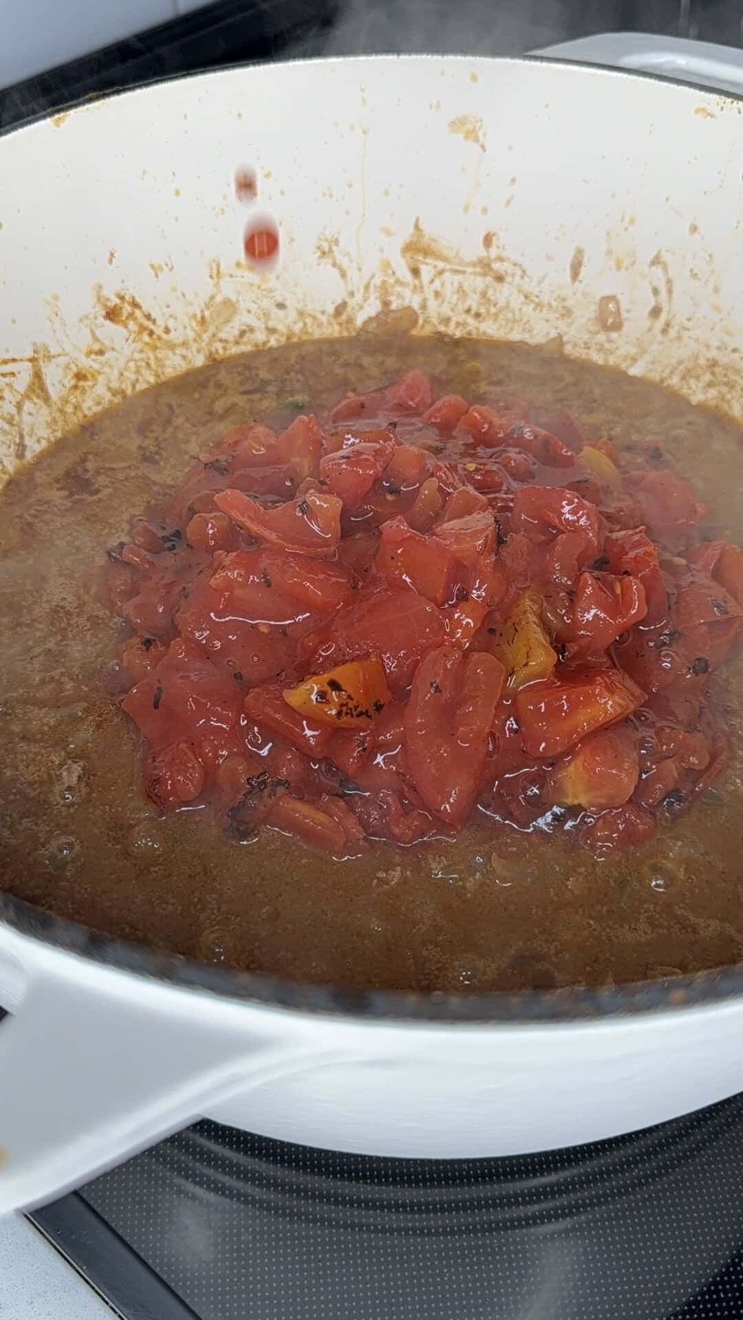 Chopped tomatoes simmer in a white pot with a brown, spiced sauce, creating steam as they cook on a stove.