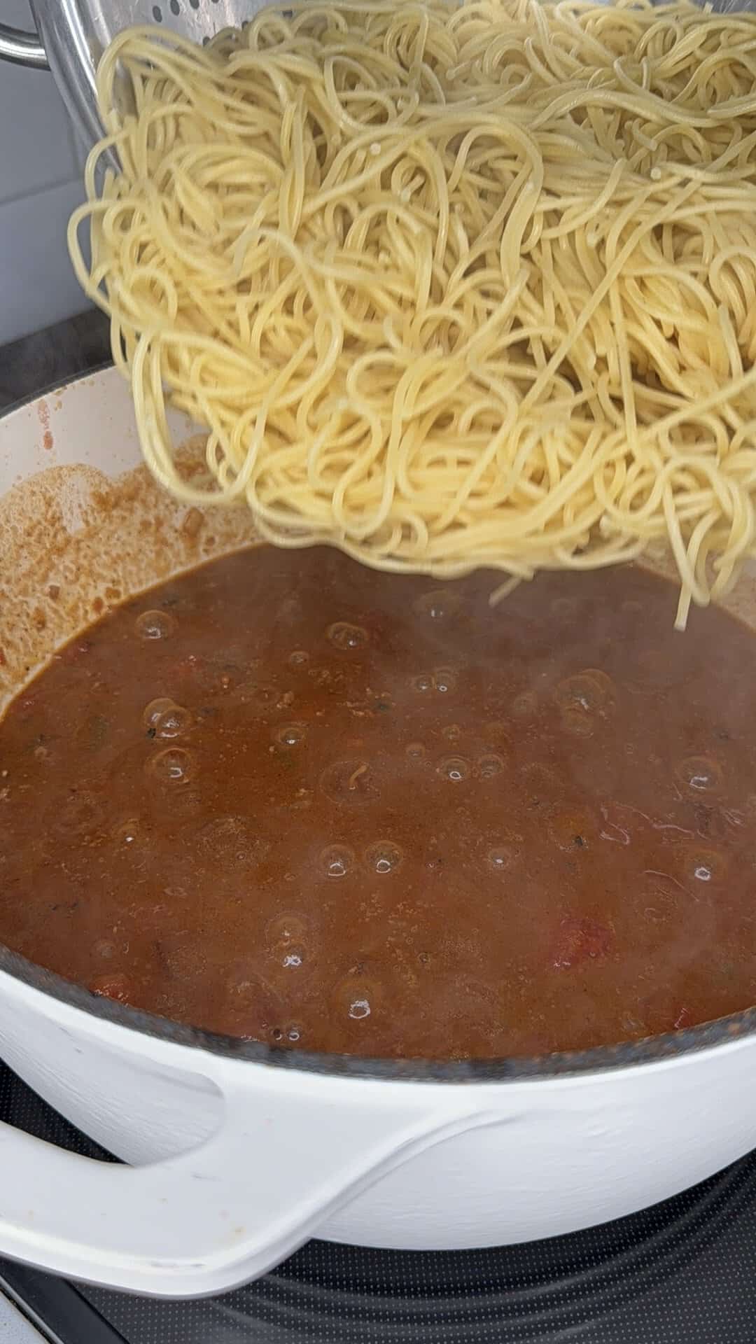 Cooked spaghetti is being poured from a colander into a white pan of bubbling red tomato sauce on a stovetop.