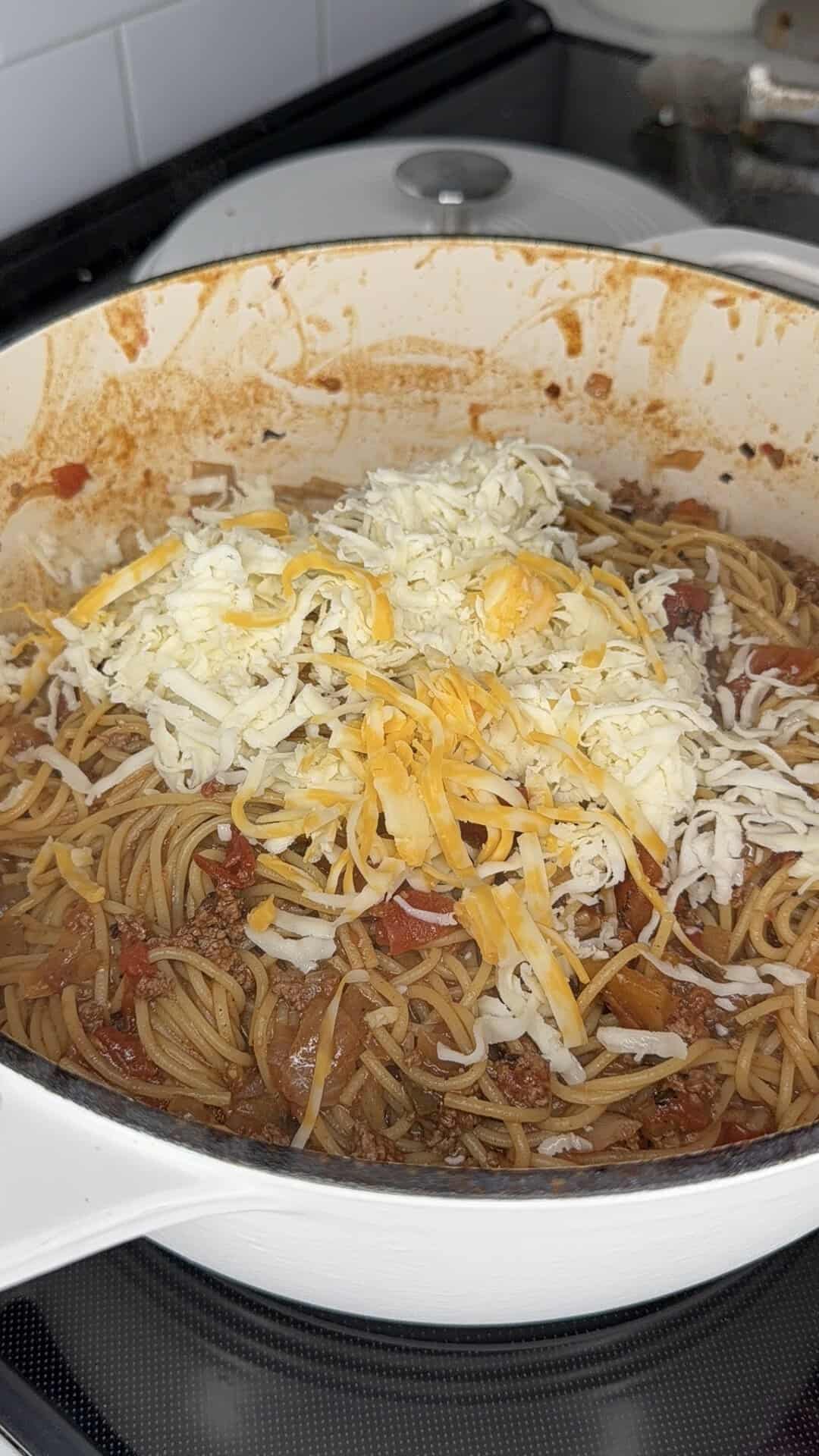 A pot of cooked spaghetti with meat sauce is topped with a pile of shredded yellow and white cheese, ready to be mixed, on a stovetop.