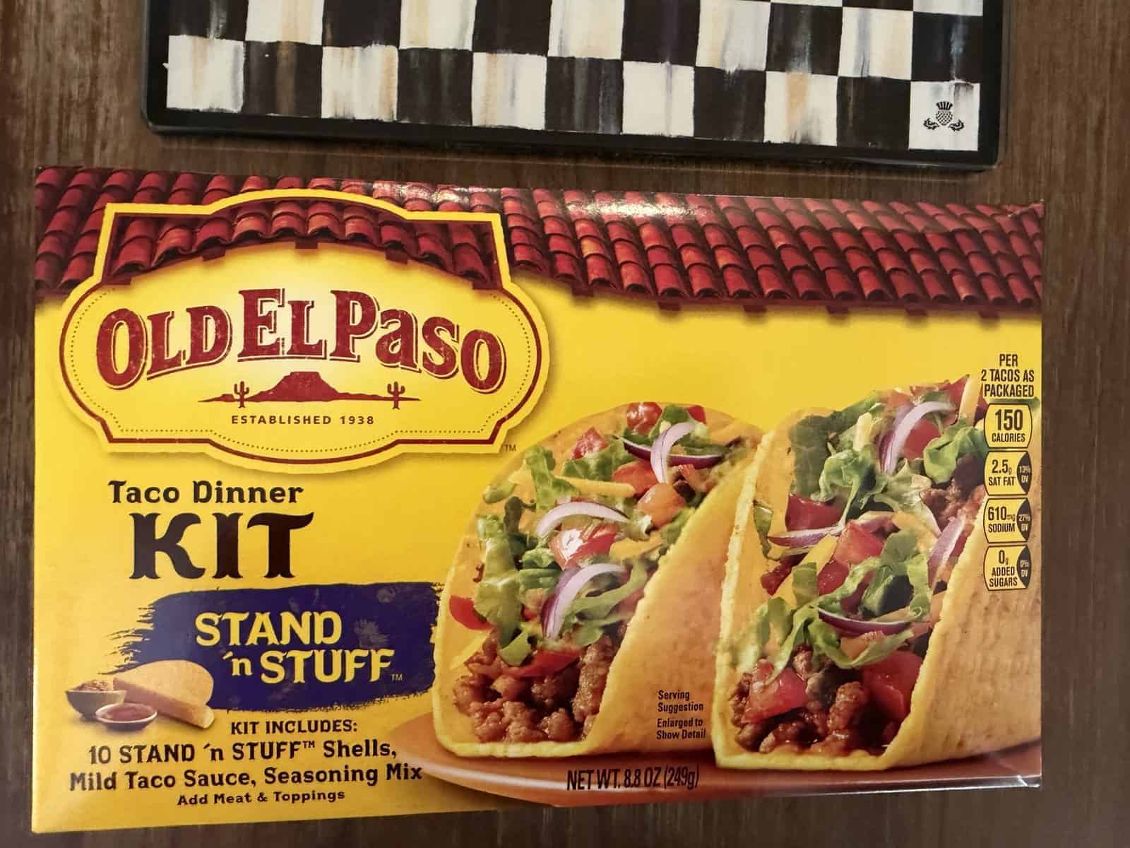 Old El Paso Taco Dinner Kit box on a table, showing two tacos filled with meat, lettuce, tomato, and cheese. Text highlights Stand n Stuff shells and includes nutrition facts on the front.