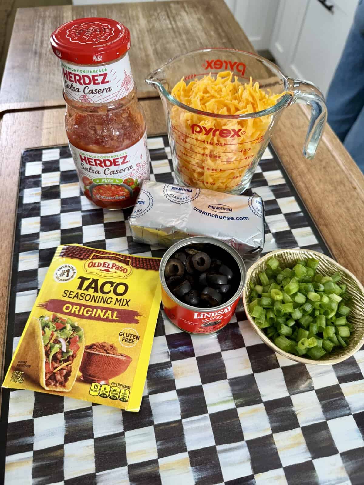 Ingredients for a dip: shredded cheese in a measuring cup, two jars of salsa, cream cheese, taco seasoning packet, sliced black olives, and chopped green onions on a checkered surface.