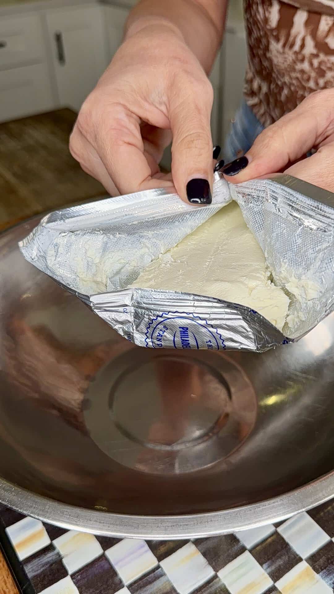 A person opens a foil-wrapped block of cream cheese over a large empty metal mixing bowl on a kitchen counter.