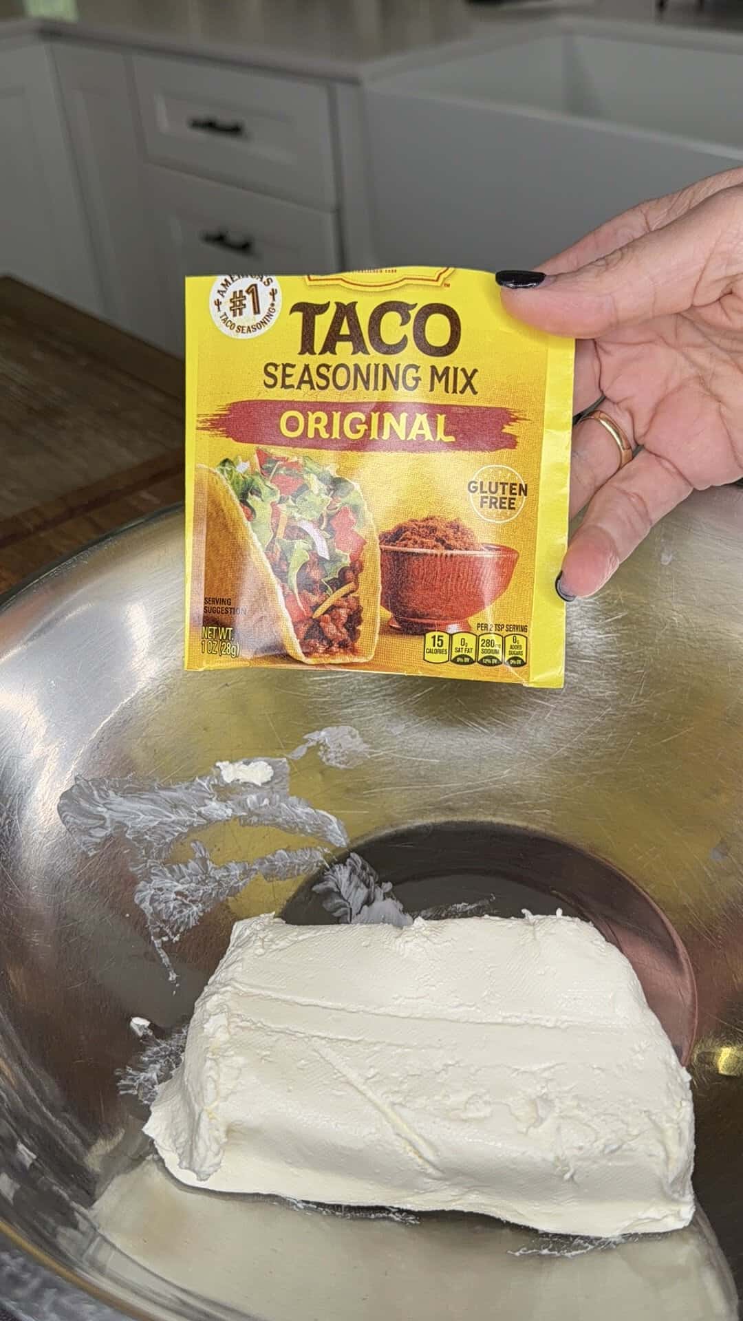 A hand holds a packet of taco seasoning mix above a metal bowl containing a block of cream cheese, with a kitchen visible in the background.