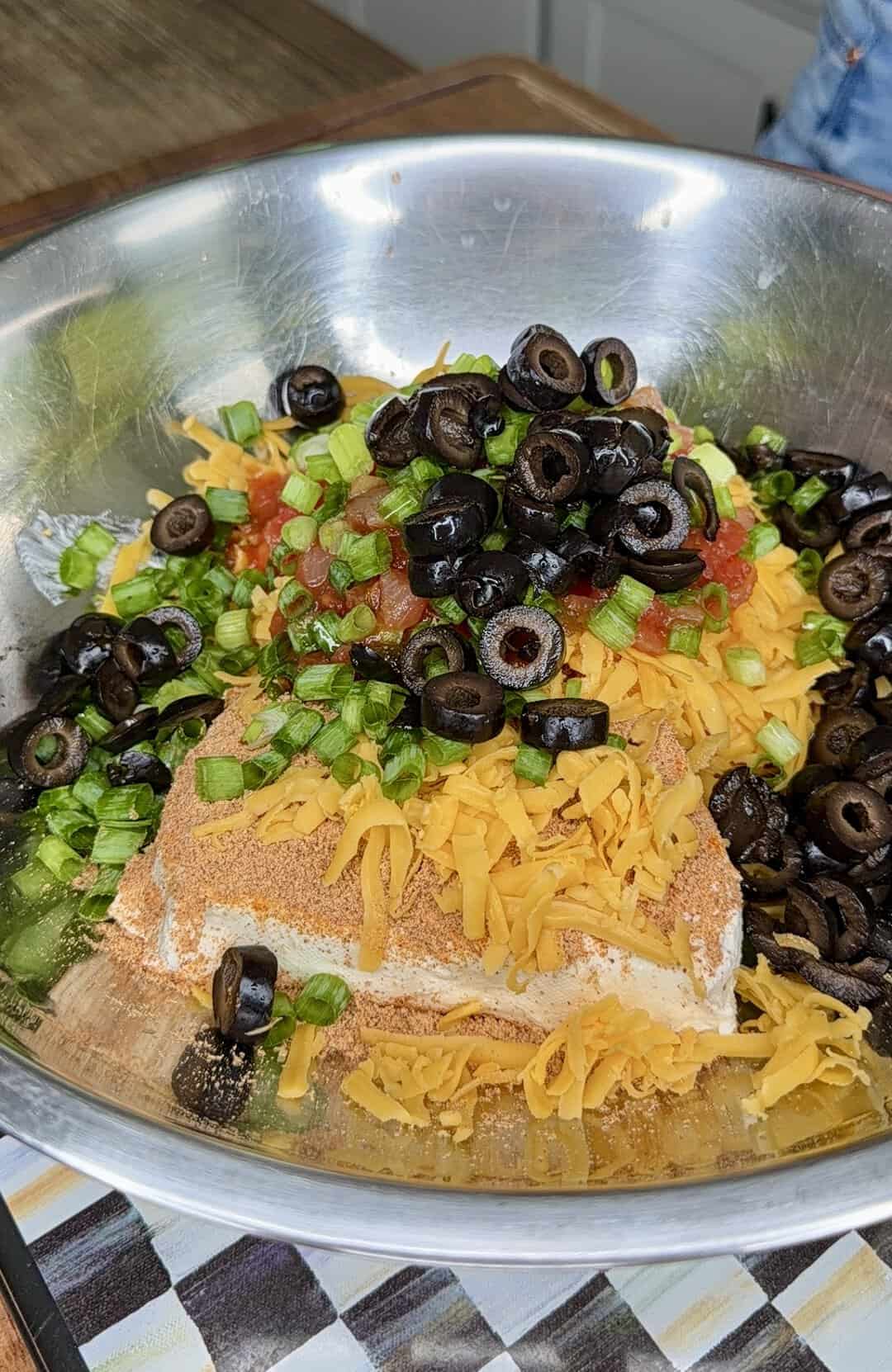 A stainless steel bowl filled with shredded cheddar cheese, chopped green onions, sliced black olives, cream cheese, salsa, and taco seasoning, ready to be mixed.