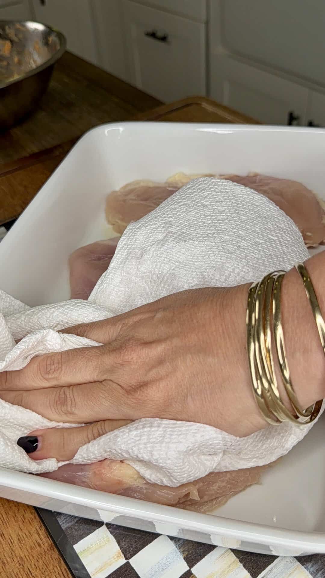 A hand wearing gold bangles pats raw chicken breasts dry with a paper towel in a white dish on a kitchen counter.