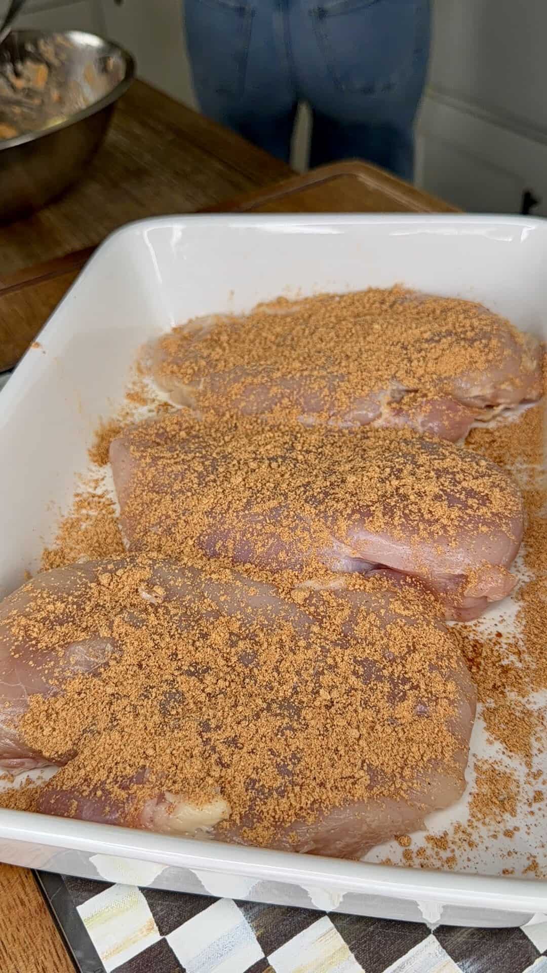 Three raw chicken breasts coated with a reddish-brown spice rub are arranged in a white baking dish on a wooden counter. A metal bowl and a person in blue jeans are in the background.