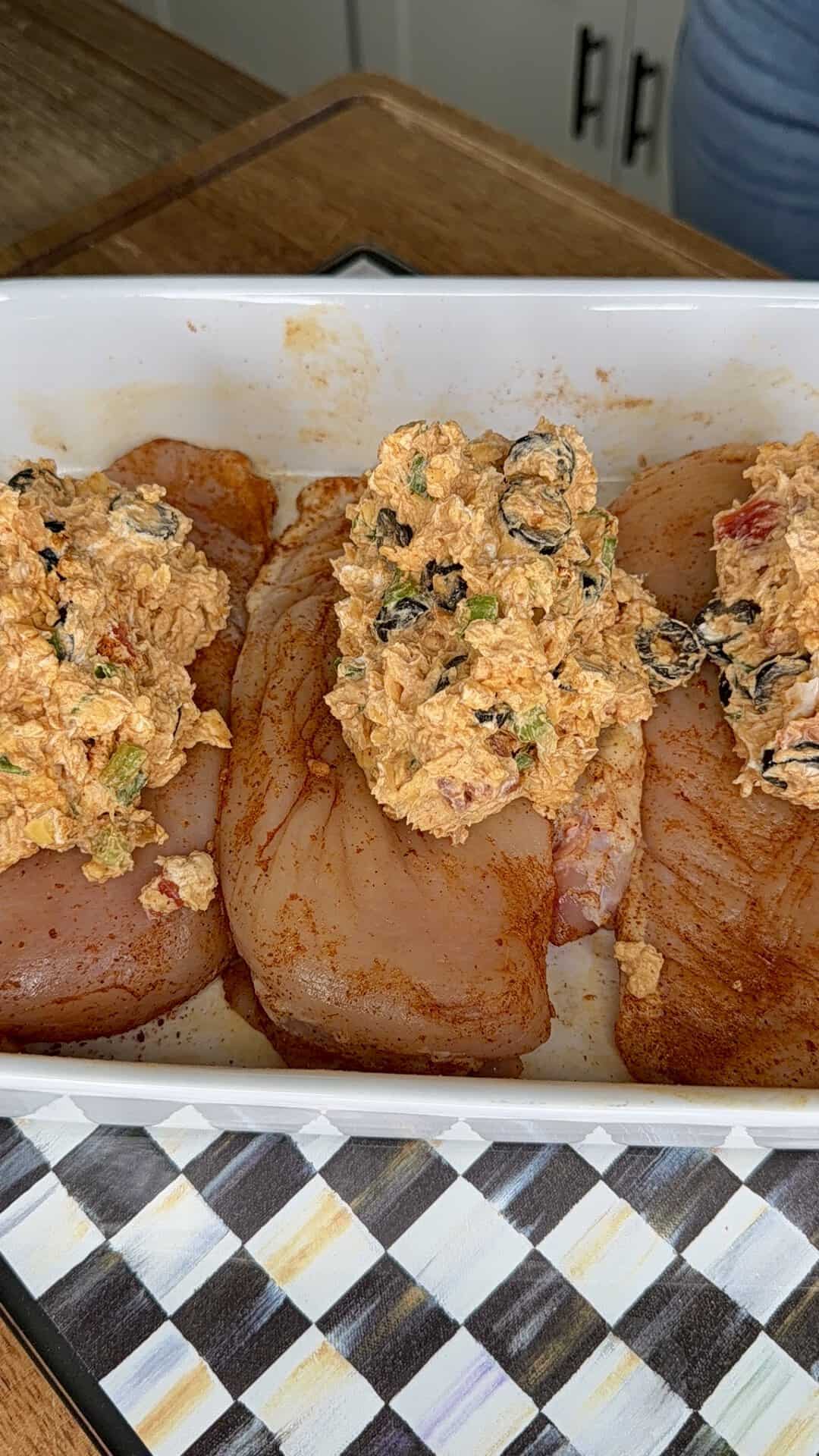 Three raw, seasoned chicken breasts in a white baking dish are topped with a mixture containing cream cheese, herbs, black olives, and diced vegetables, placed on a checkered towel on a wooden counter.