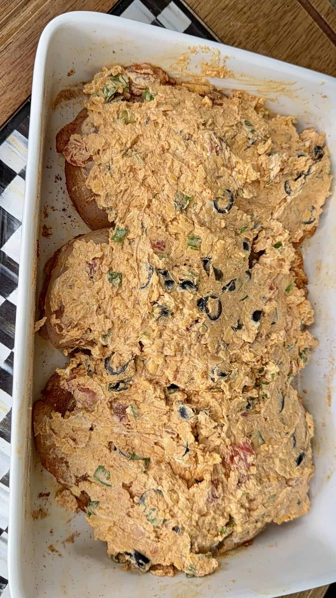 A white baking dish contains bread slices topped with a creamy, orange spread mixed with vegetables and black olives. The dish sits on a wooden table with a checkered napkin partially visible underneath.