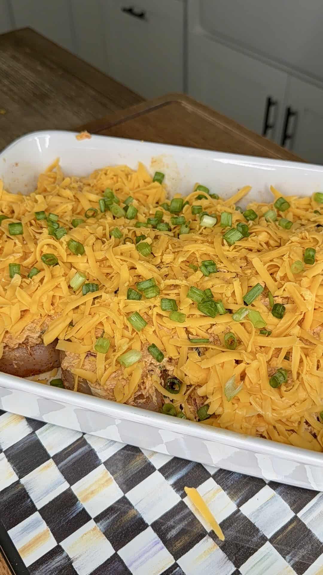 A white casserole dish filled with sliced bread topped with shredded cheddar cheese and chopped green onions, sitting on a patterned surface.