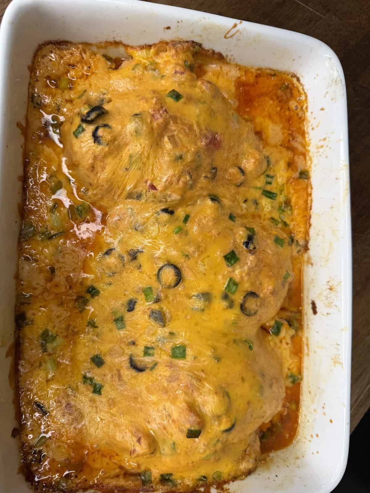 A baked casserole topped with melted cheddar cheese, sliced black olives, and chopped green onions in a white dish. Orange-colored sauce is visible around the edges.
