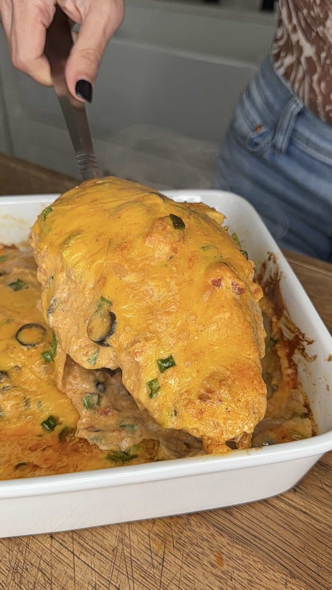 A person serves a cheesy, golden-baked casserole with green onions from a white baking dish using a spatula.
