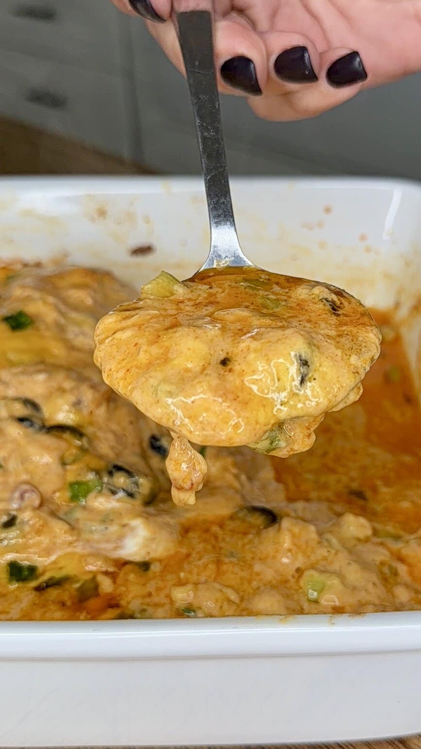 A spoon holding a cheesy, saucy casserole mixture with visible vegetables, lifted from a white dish. A hand with dark nail polish holds the spoon.