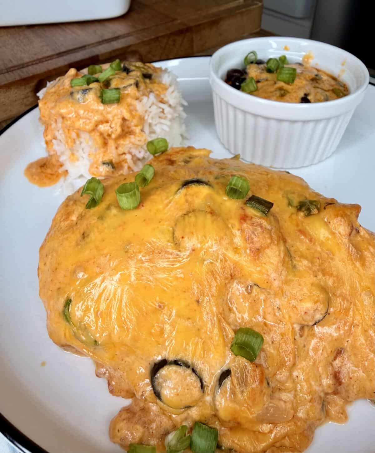 A plate with cheesy chicken casserole topped with green onions, served alongside a mound of white rice and a small ramekin of more casserole, garnished with green onions.