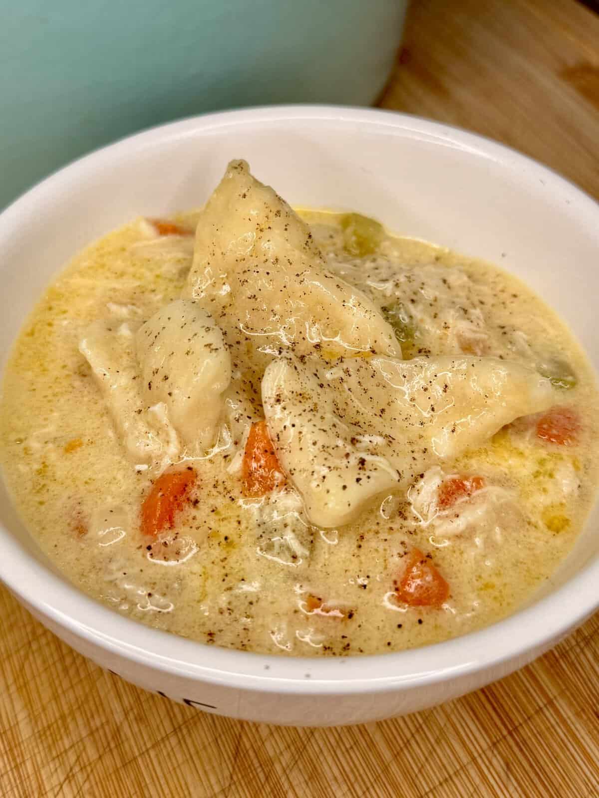 A white bowl filled with creamy chicken and dumplings soup, featuring chunks of chicken, diced carrots, and celery, topped with black pepper. This easy family dinner sits on a wooden surface.